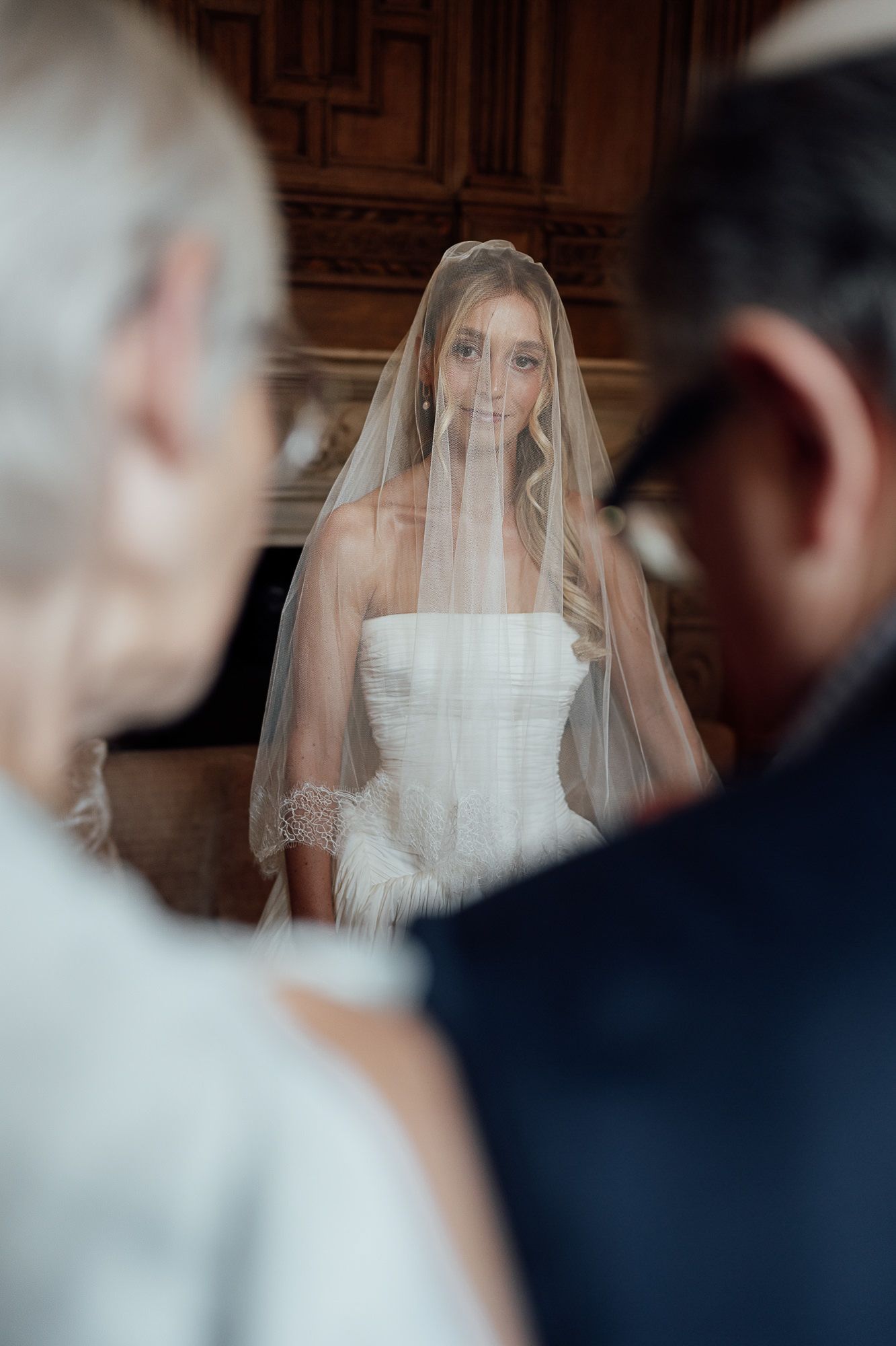Bride in white strapless gown and veil smiles at two guests during an elegant indoor wedding ceremony.