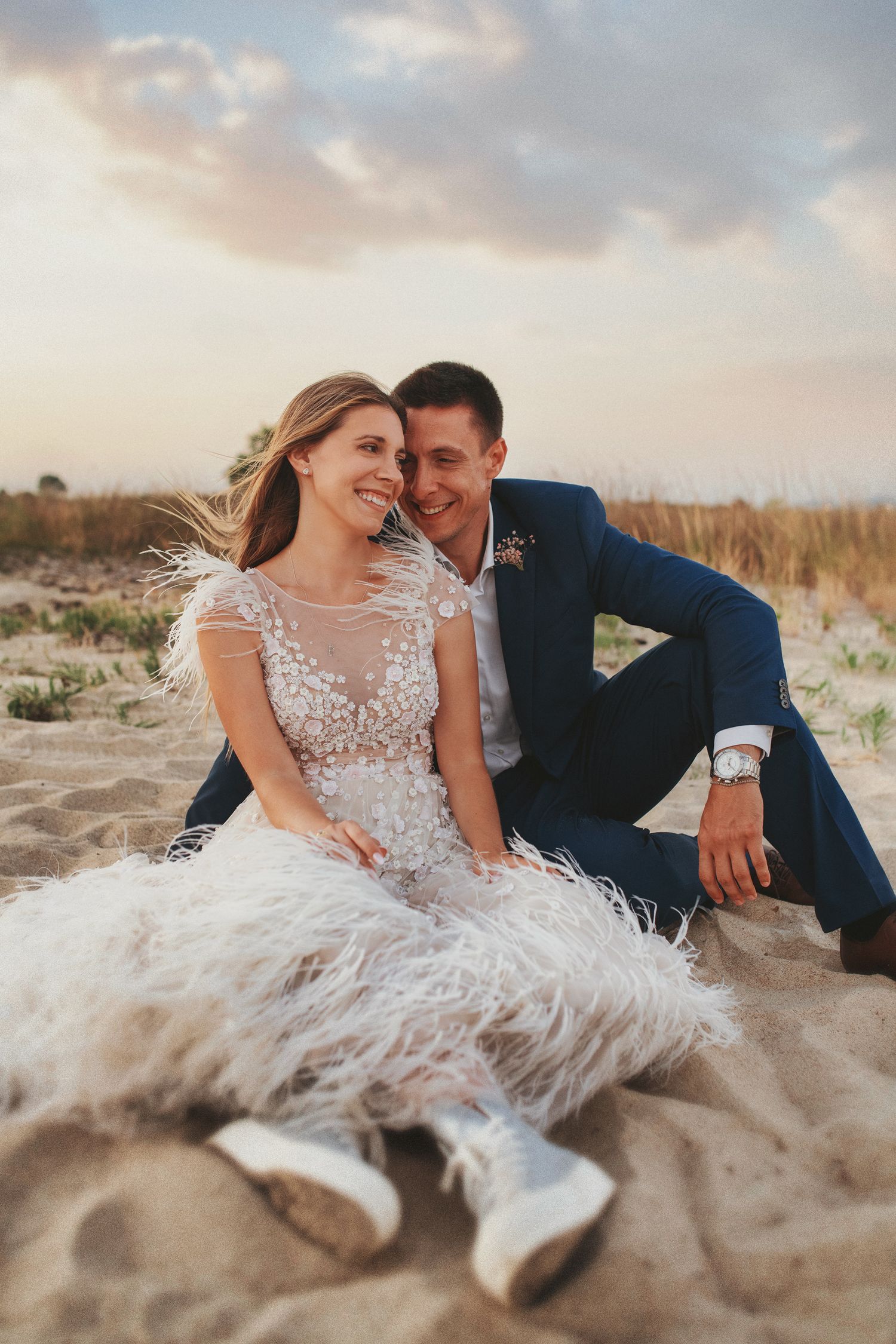 Candid and joyful wedding photography with a bride and groom laughing while sitting on a sandy beach at sunset.