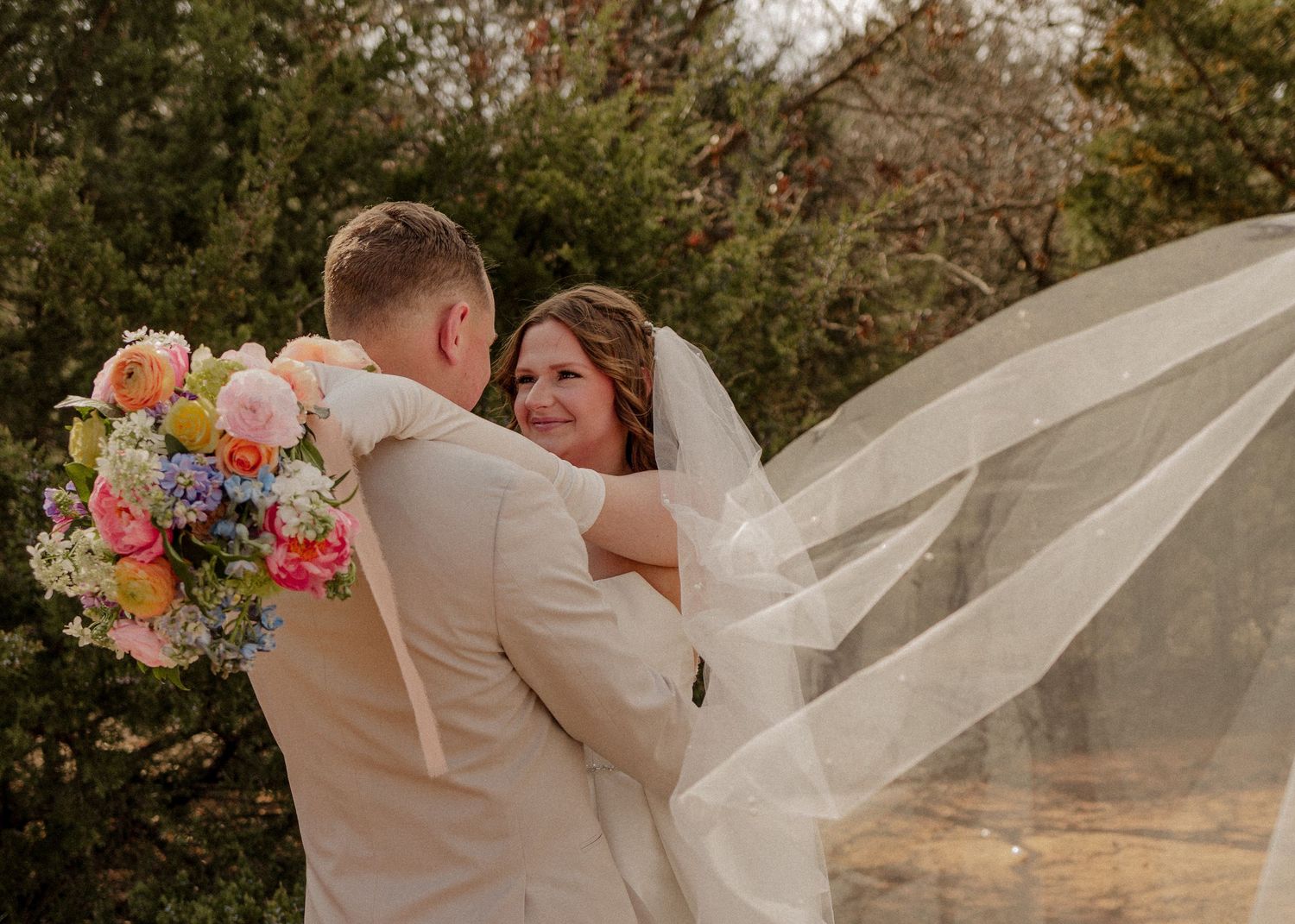 Bride smiling at groom with colorful bouquet and flowing veil in garden. Esperanza Ranch bride and groom portraits in Oklahoma