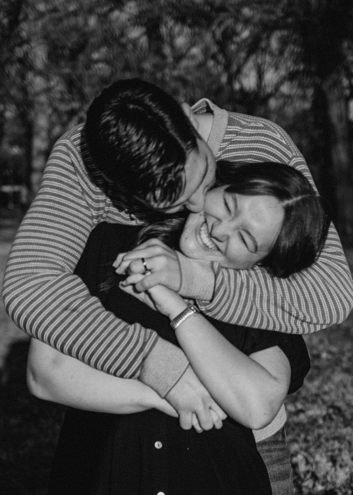 A black and white photo of a man hugging a laughing woman from behind outdoors. Oklahoma City couple photoshoot.