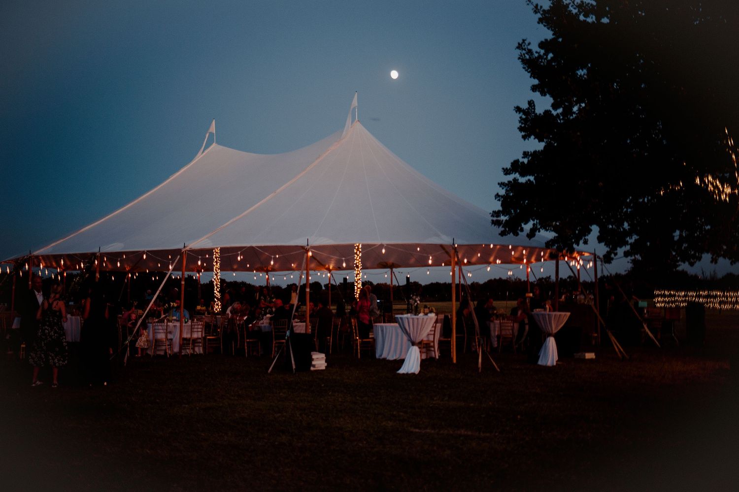 Elegant wedding tent glowing with string lights under a moonlit night sky at an outdoor reception. Oklahoma Spring wedding on a family farm