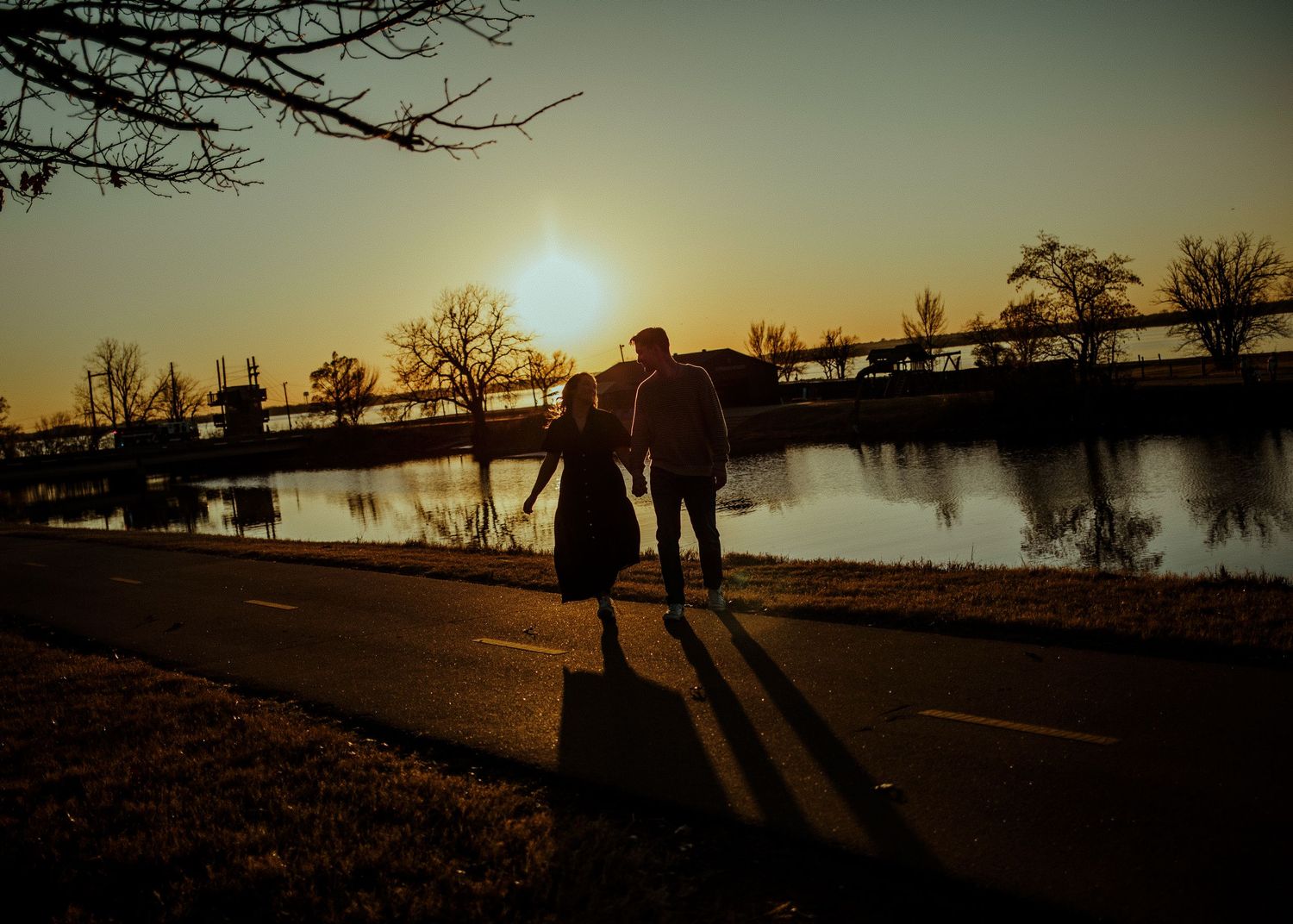 A couple holds hands in silhouette while walking beside a river at sunset. Oklahoma City couple photoshoot.