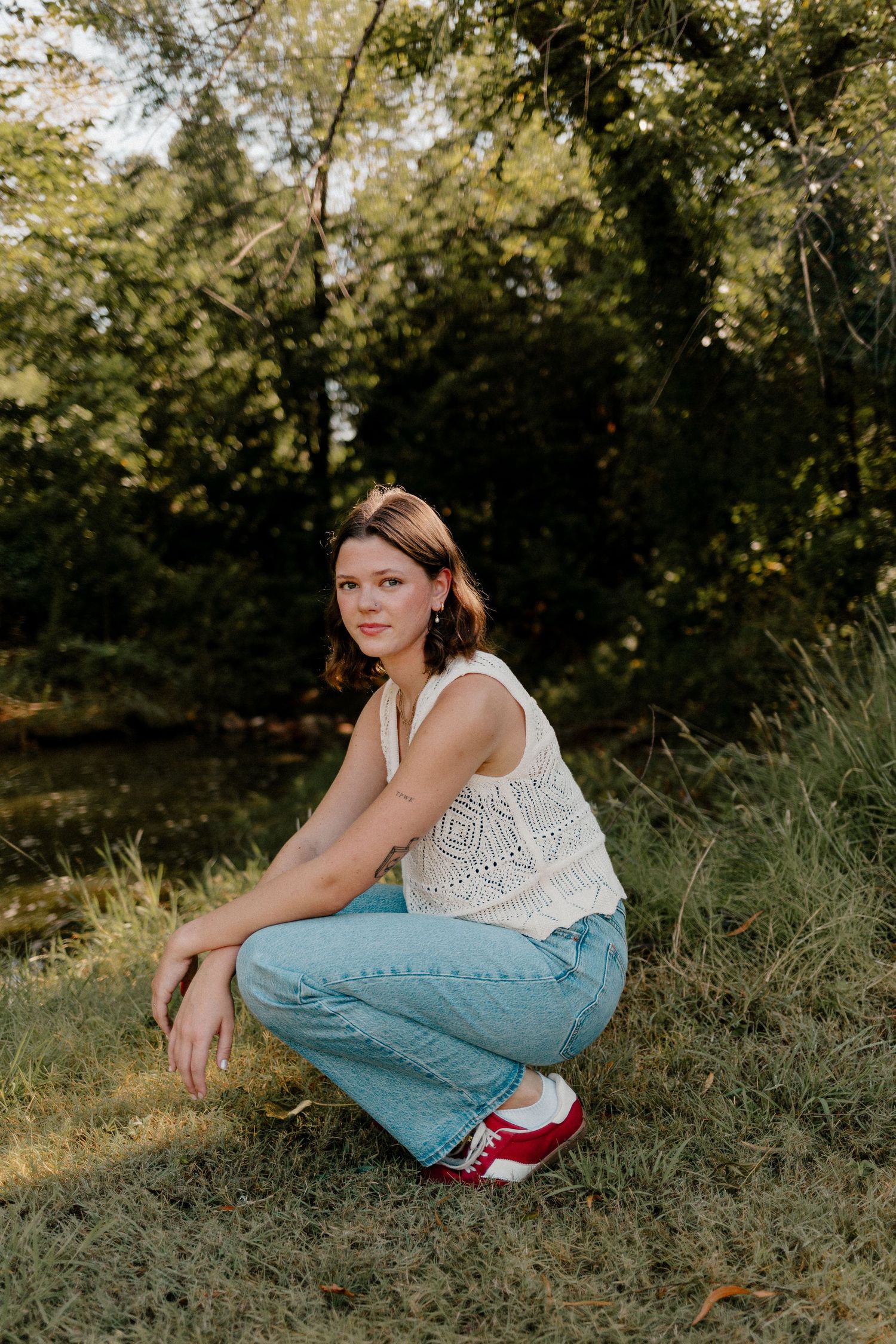 Young woman in white crochet top and jeans crouching on grass in a lush green park. Oklahoma City senior pictures