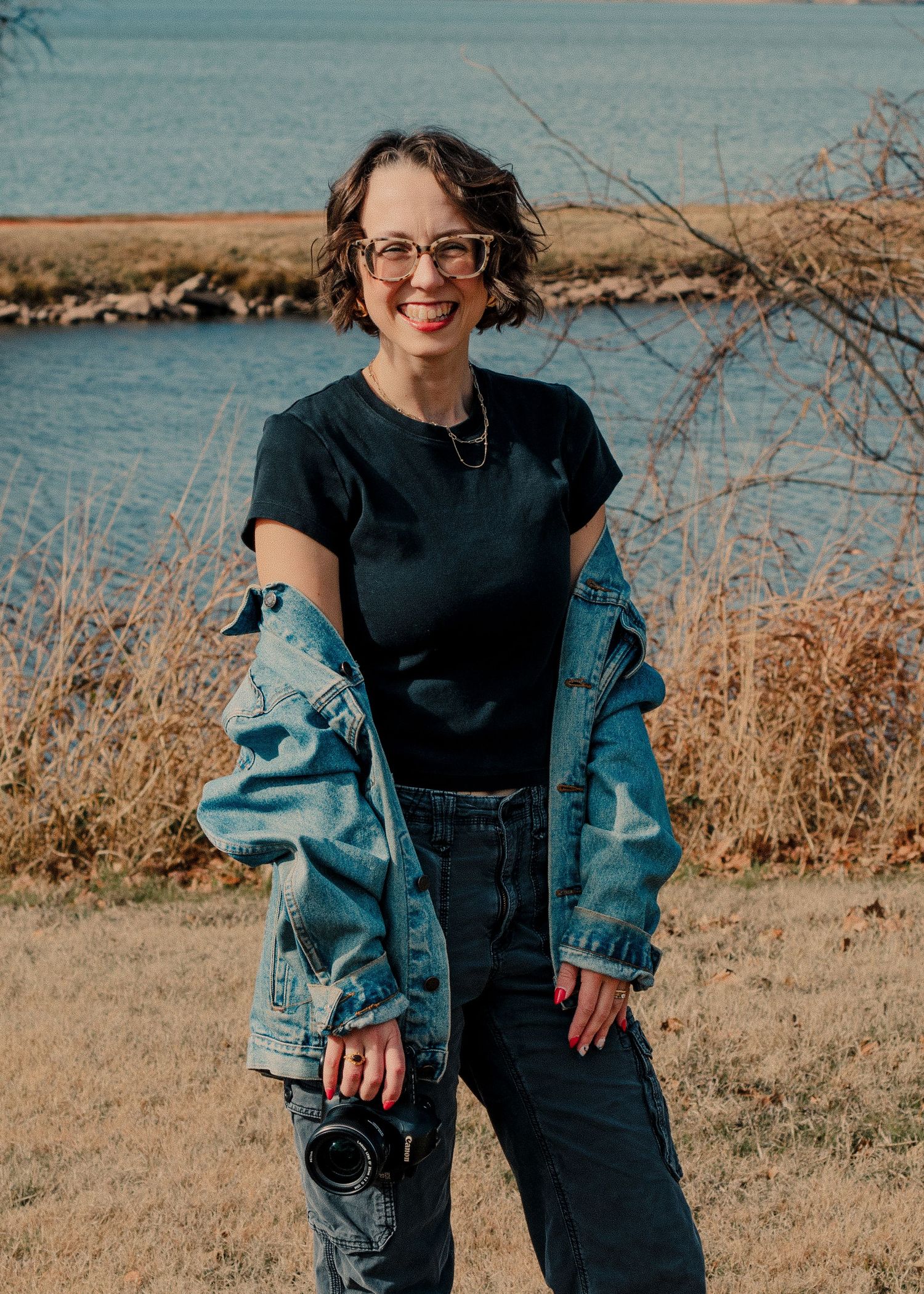 Female photographer smiling by lakeside holding camera in denim jacket. Addi Oklahoma based wedding photographer