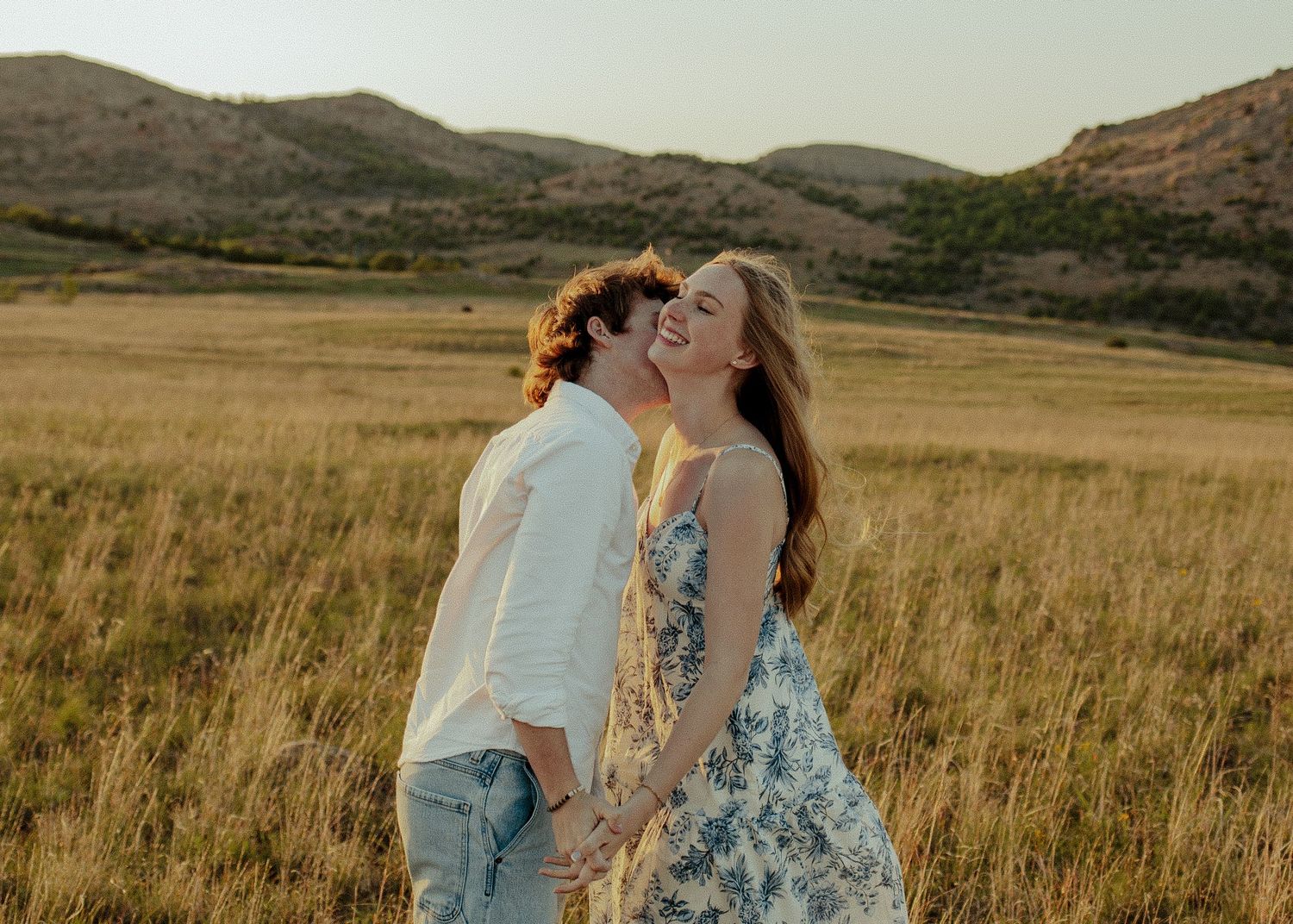 Couple sharing a joyful kiss in a sunlit meadow with mountain backdrop. Wichita mountains engagement photoshoot