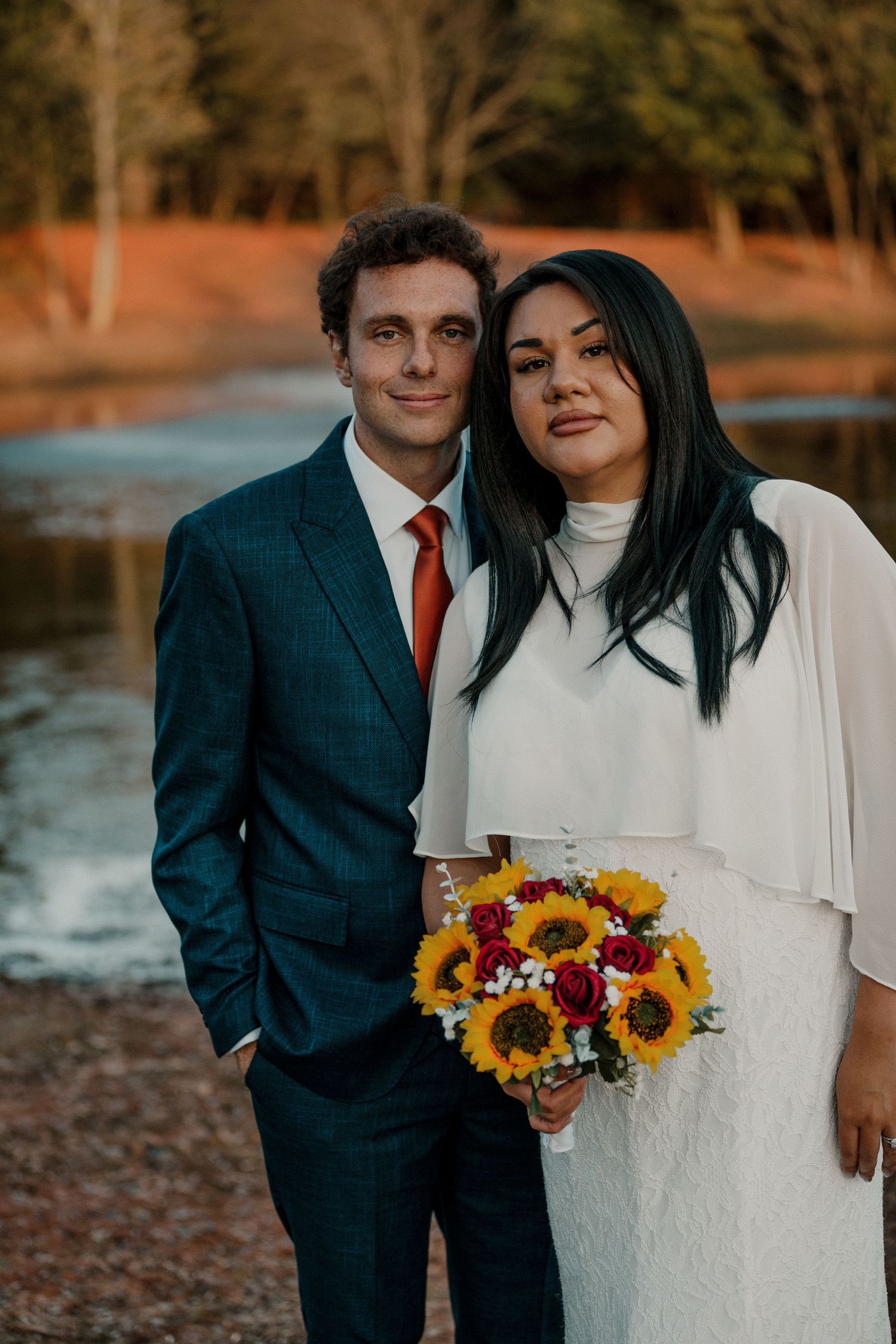 Newlyweds posing by a lake, bride holding sunflower bouquet, groom in navy suit. Oklahoma City intimate fall wedding.