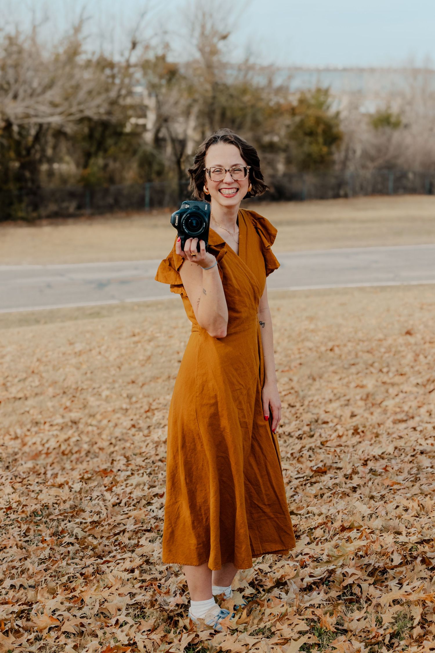 Female photographer in rust dress holding camera among autumn fallen leaves. Addi Suenram Oklahoma based wedding photographer