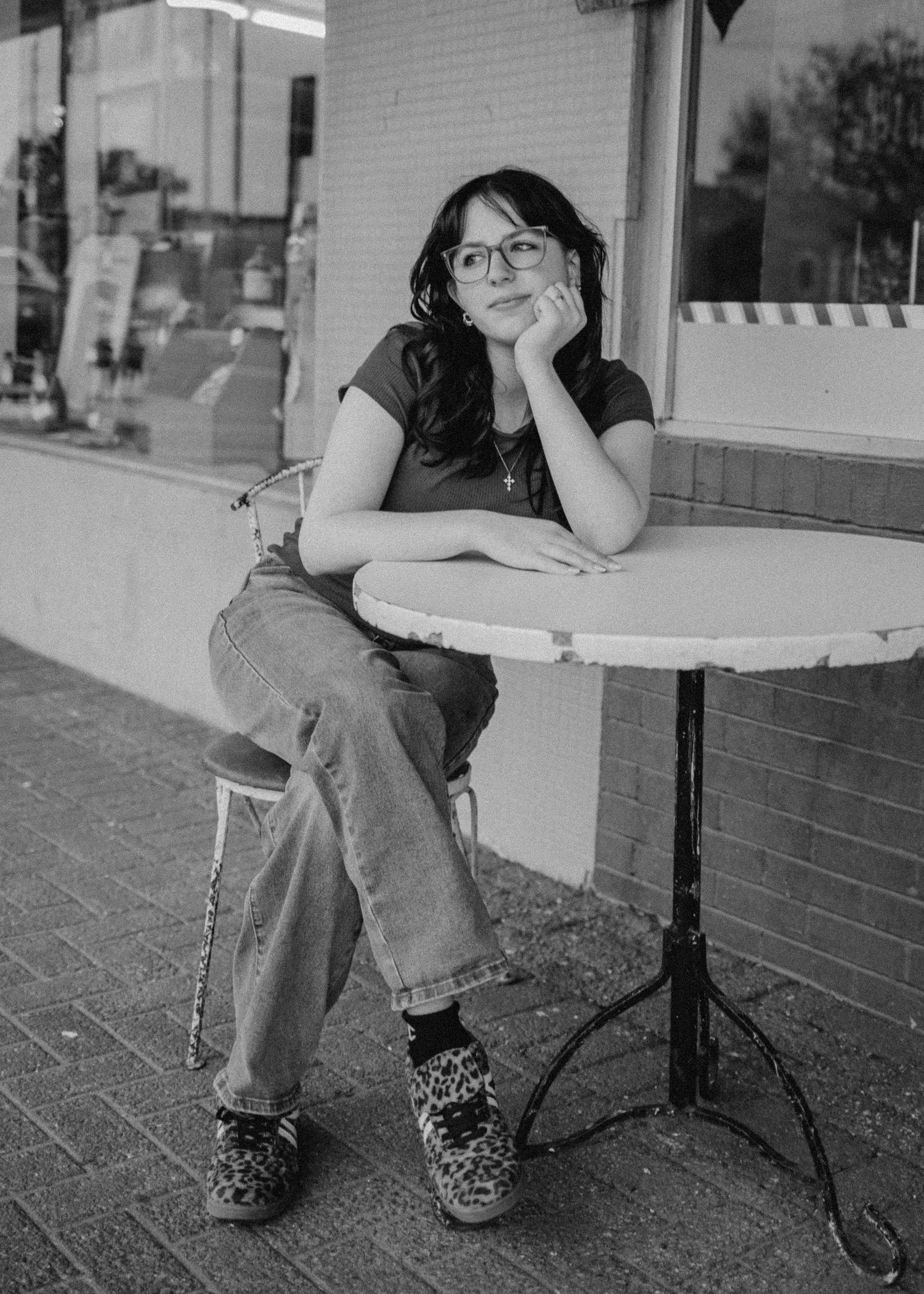 Black and white senior portrait of a young woman sitting at an outdoor cafe table with glasses. Bethany Oklahoma photoshoot