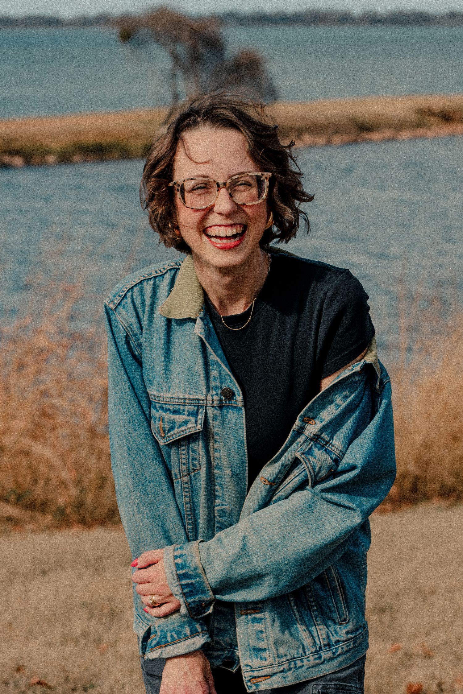 Smiling woman with glasses laughing outdoors near a lake in denim jacket. Addi Suenram Oklahoma based wedding photographer