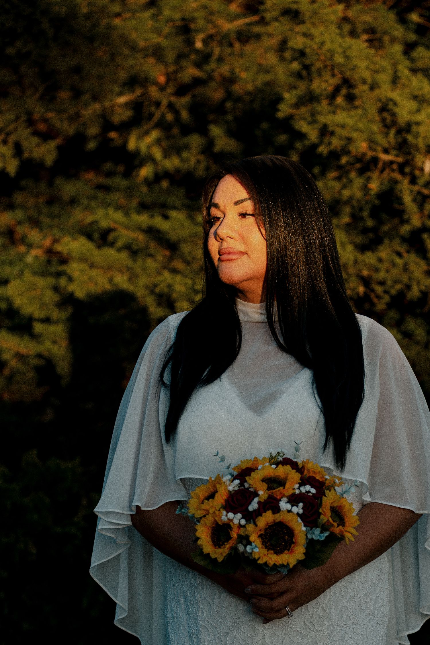 Bride in white cape dress holding sunflower bouquet with lush green trees in golden hour light. Outdoor fall wedding in Oklahoma City