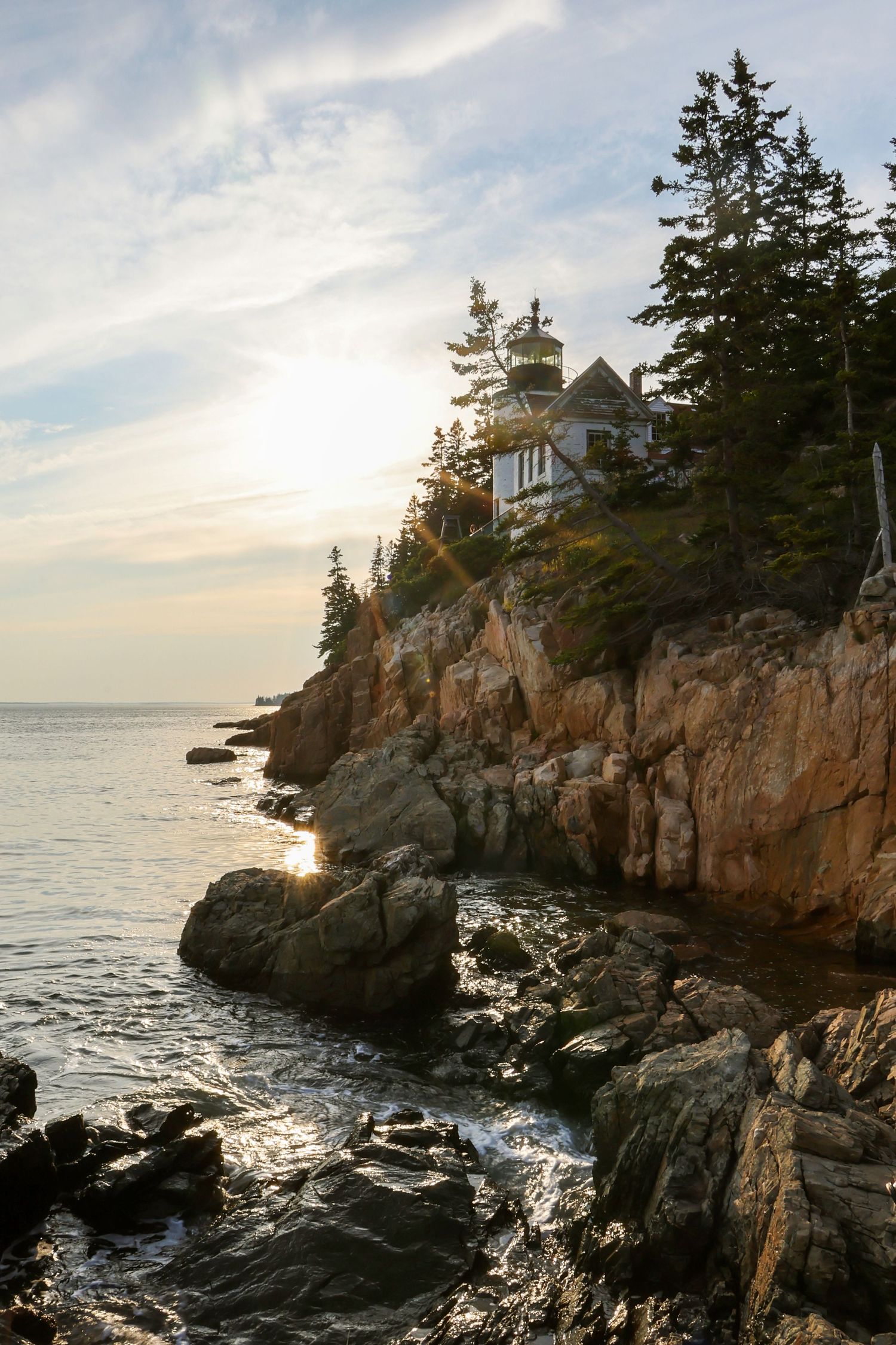 Bass Harbor Head Lighthouse sits atop rugged rocky cliffs with pine trees silhouetted against sunset sky.