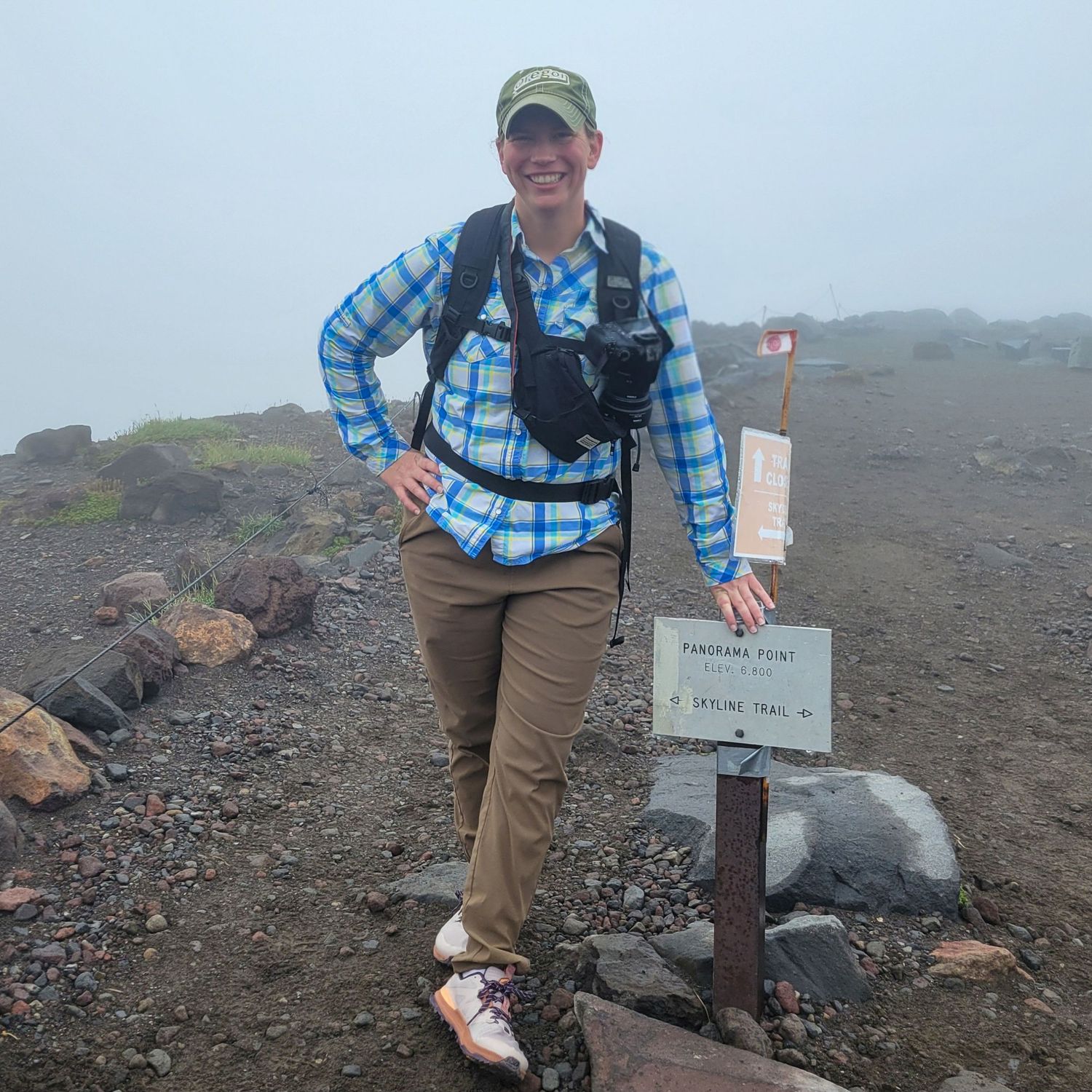 A hiker smiles at Panorama Point trail sign on a foggy volcanic mountain summit with backpack.