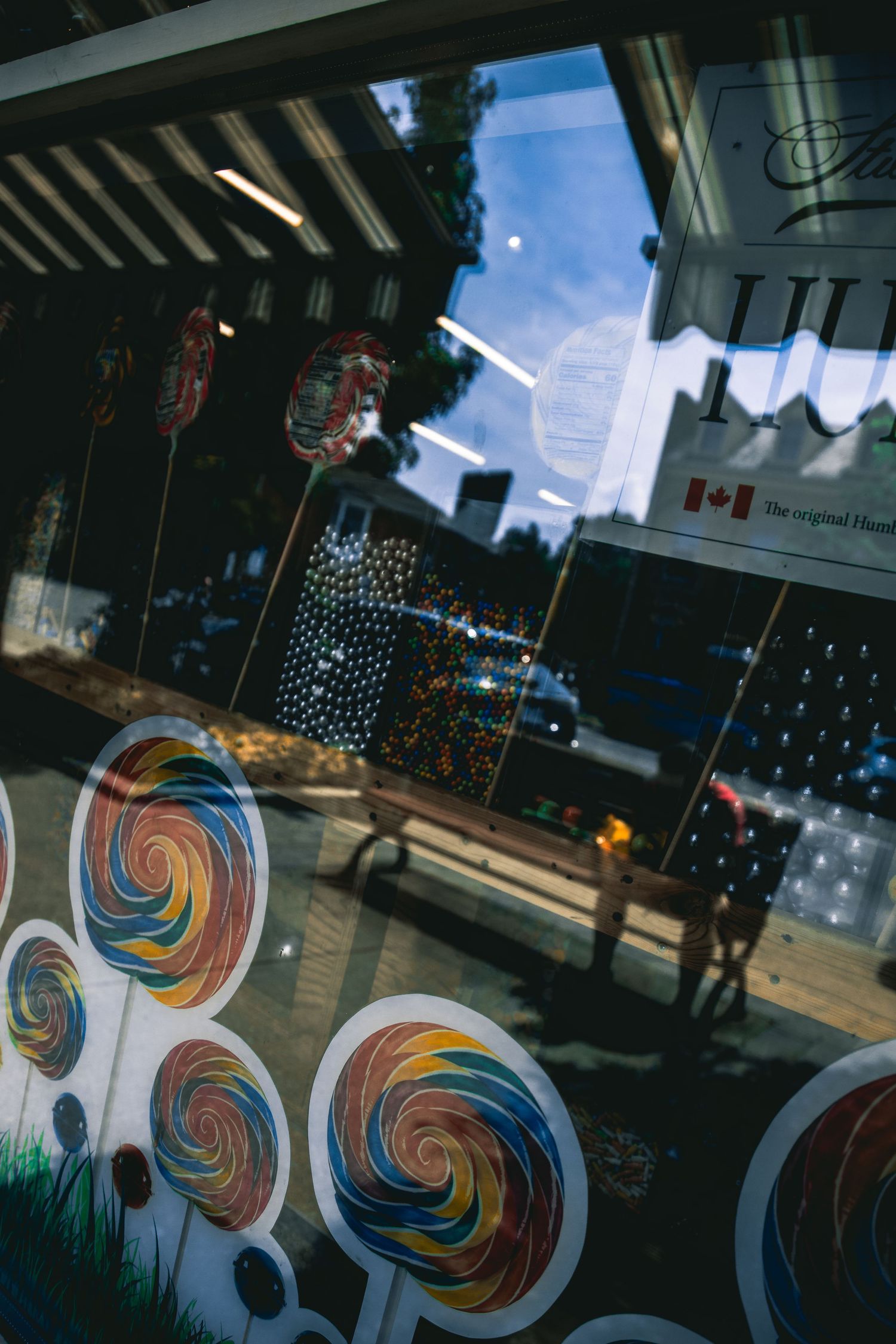 Colorful lollipop display in a candy shop window with Canadian flag reflection on glass.