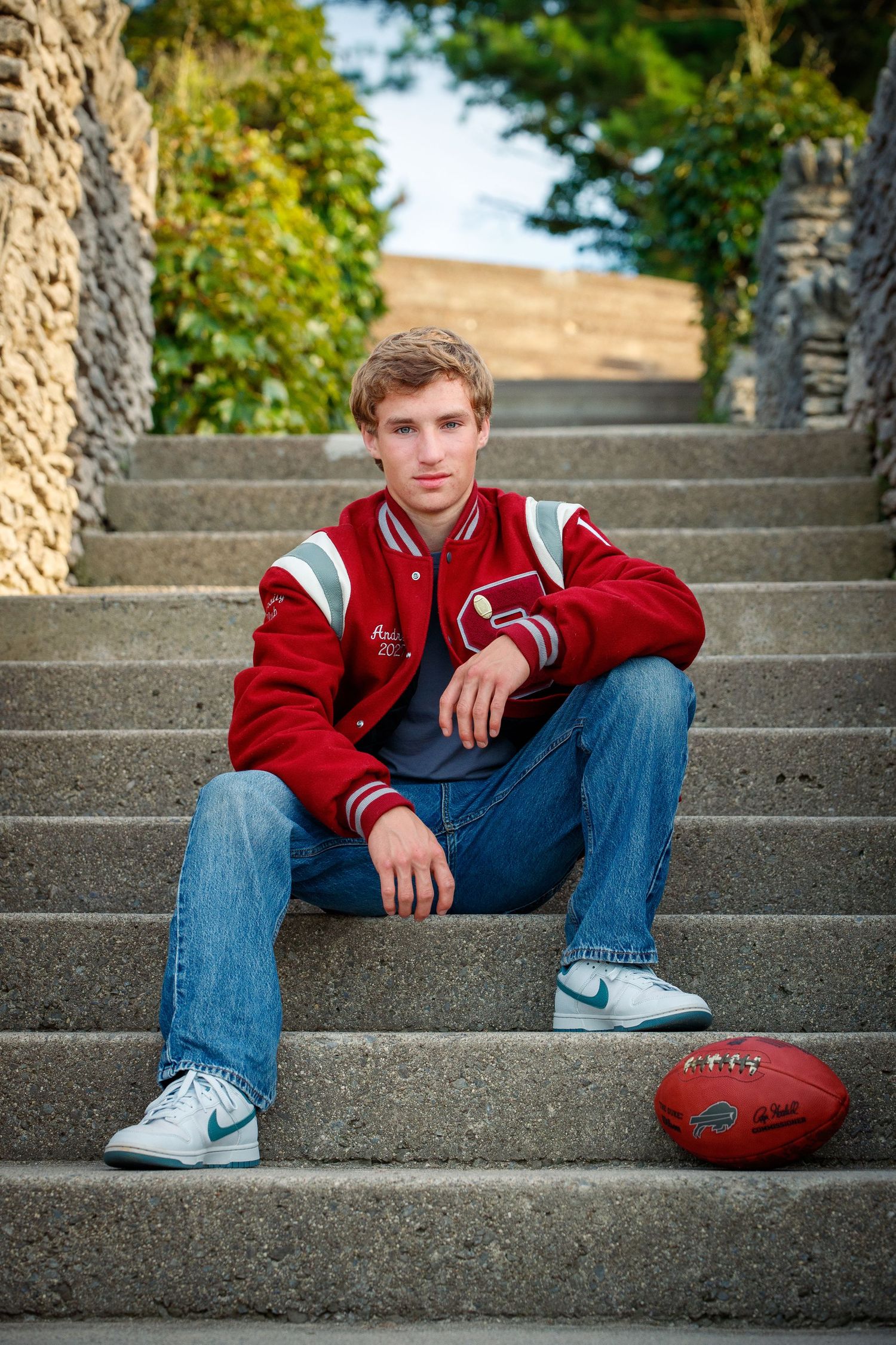 Young man in red varsity jacket and jeans sits on stone steps with a football for senior portraits.