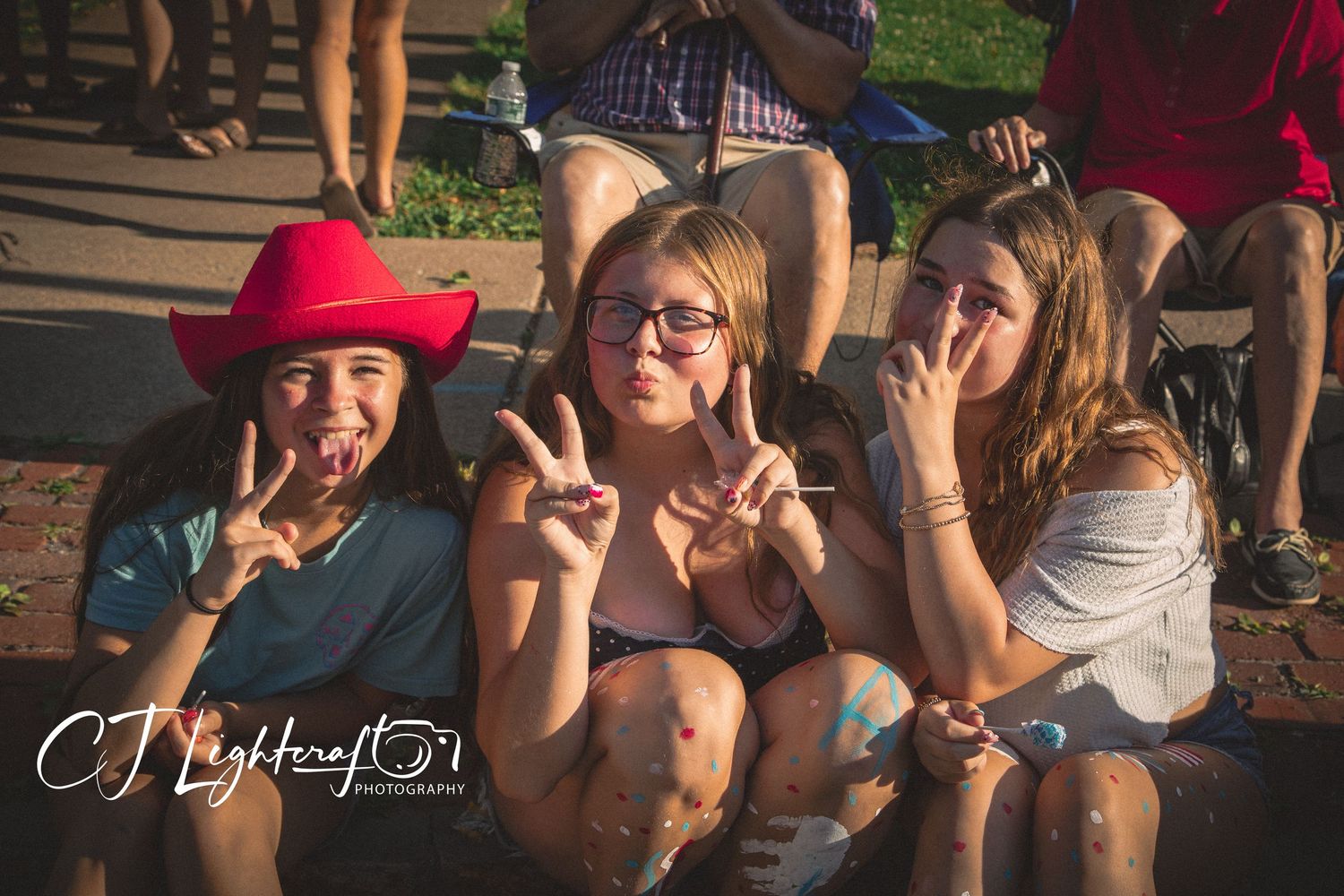 Three young women make peace signs while sitting outdoors, one wearing a red cowboy hat.