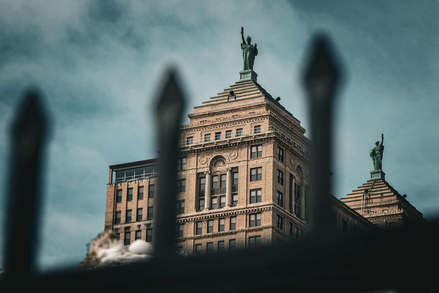 Historic brick building with green Statue of Liberty replicas on rooftop viewed through iron fence.
