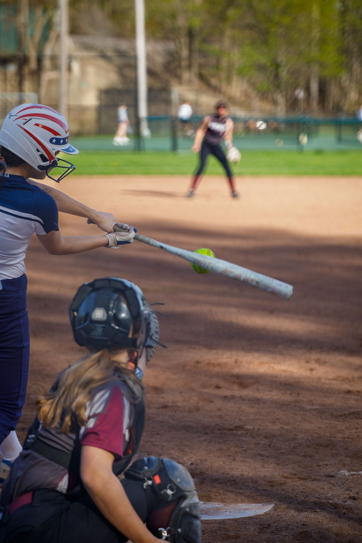 Softball batter swings at a pitch during a youth girls softball game with catcher and pitcher visible on the field.