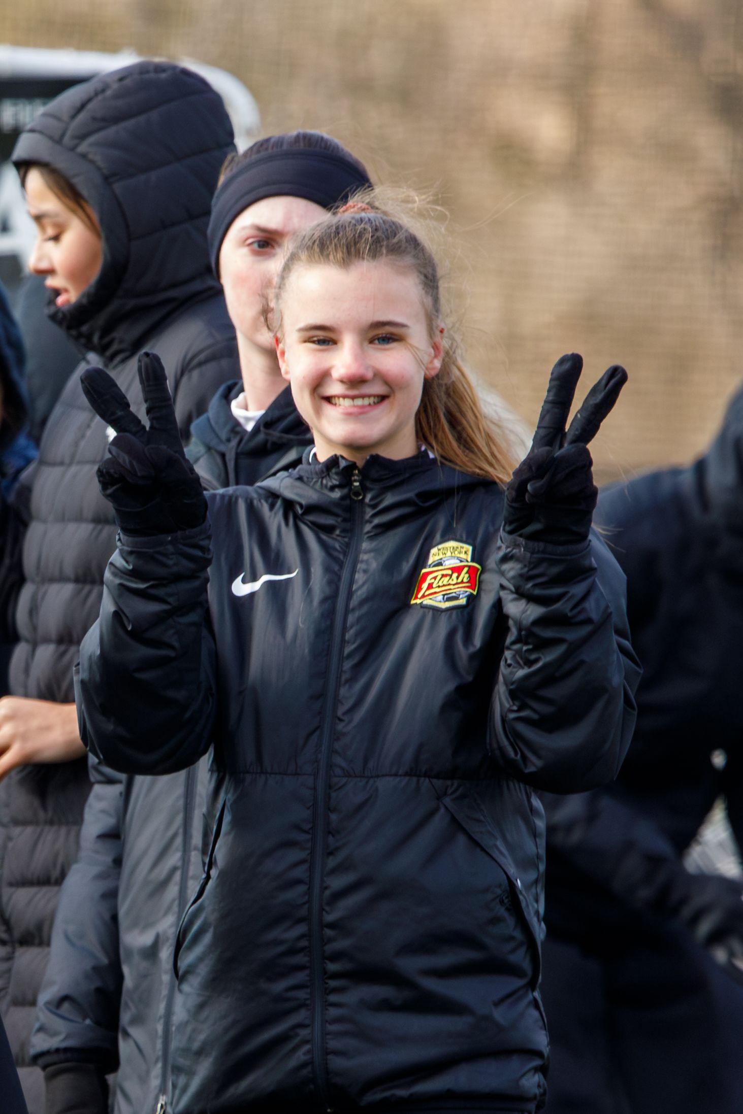 Smiling female soccer player flashes peace signs wearing a black Nike Flash Soccer jacket outdoors.