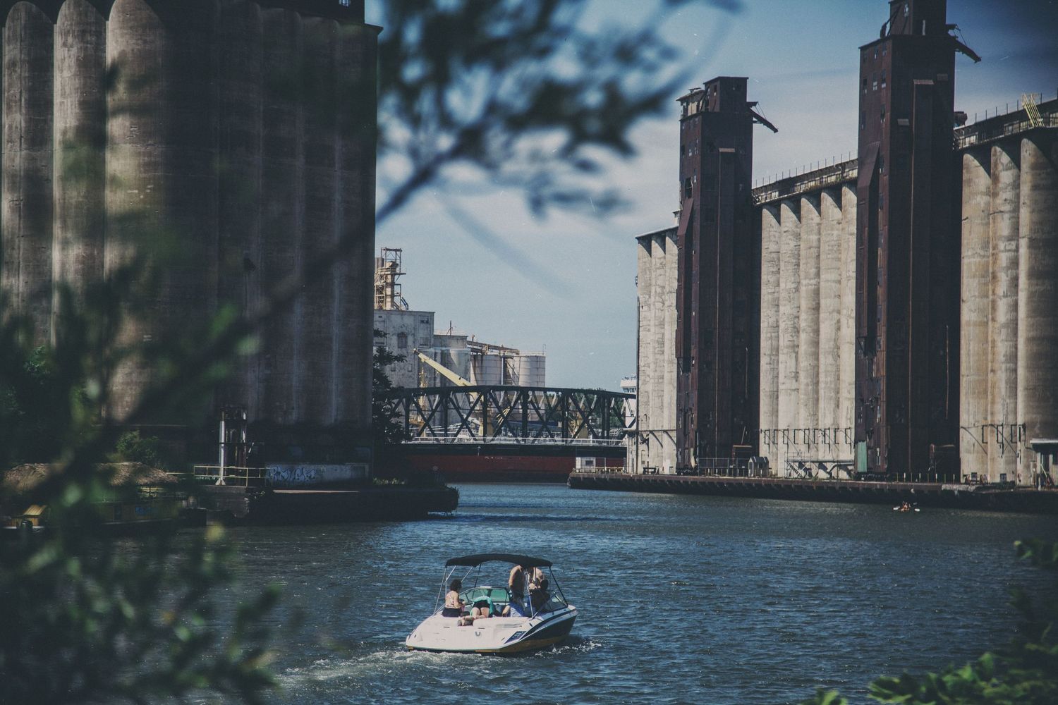 Small motorboat cruising along industrial waterway beneath bridge with large concrete grain silos in background.
