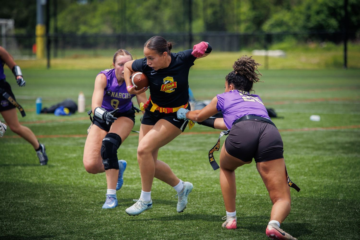 Women's flag football players compete on green turf field during outdoor tournament game.