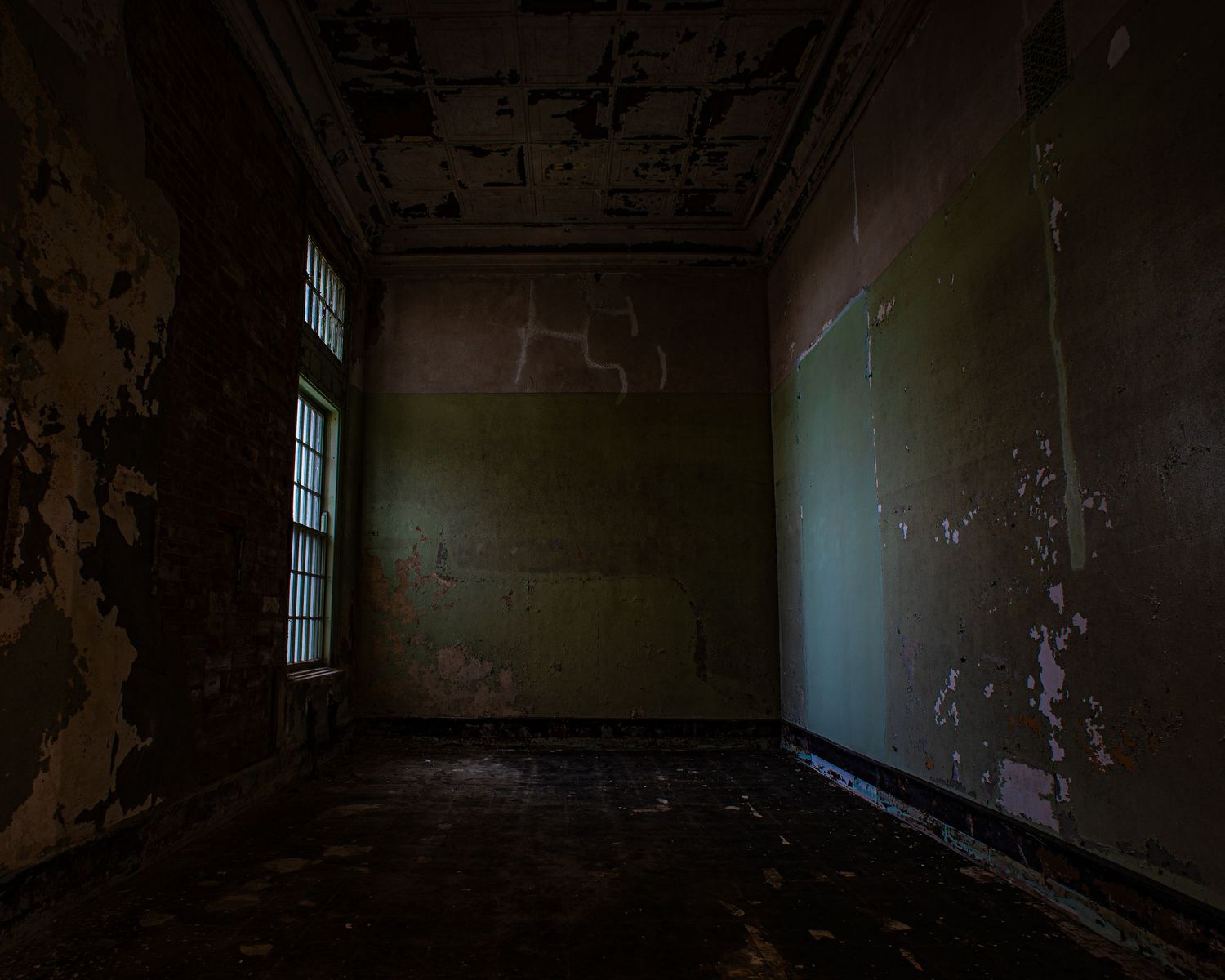Dark abandoned room with peeling paint walls, deteriorating ceiling, and dim light through barred window.