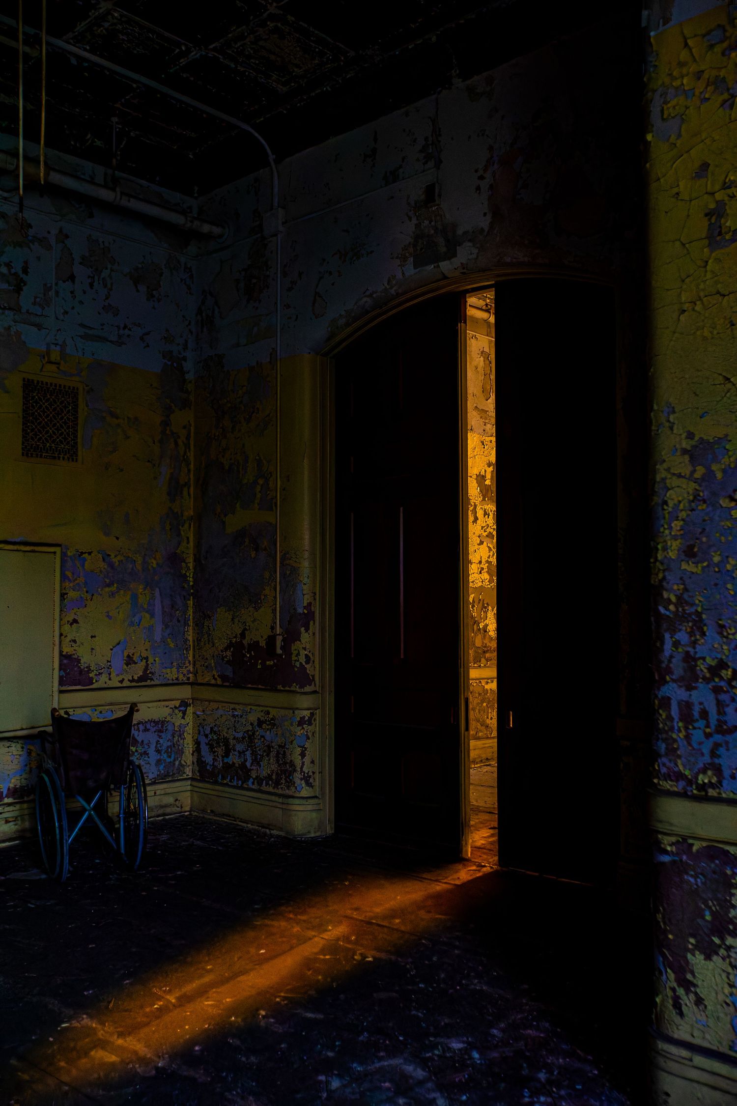Abandoned room with peeling blue walls, wheelchair, and warm light streaming through a dark doorway.