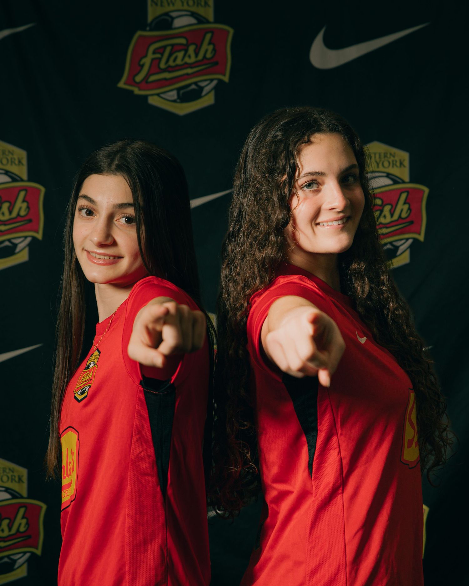 Two female soccer players in red Flash jerseys point at camera during team photo shoot.