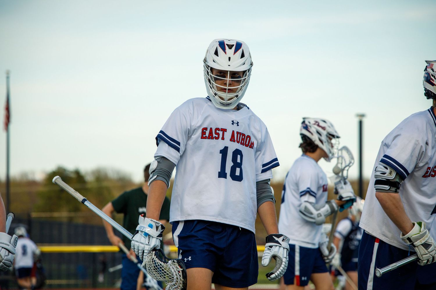 East Aurora lacrosse player #18 in white uniform holding stick on field.
