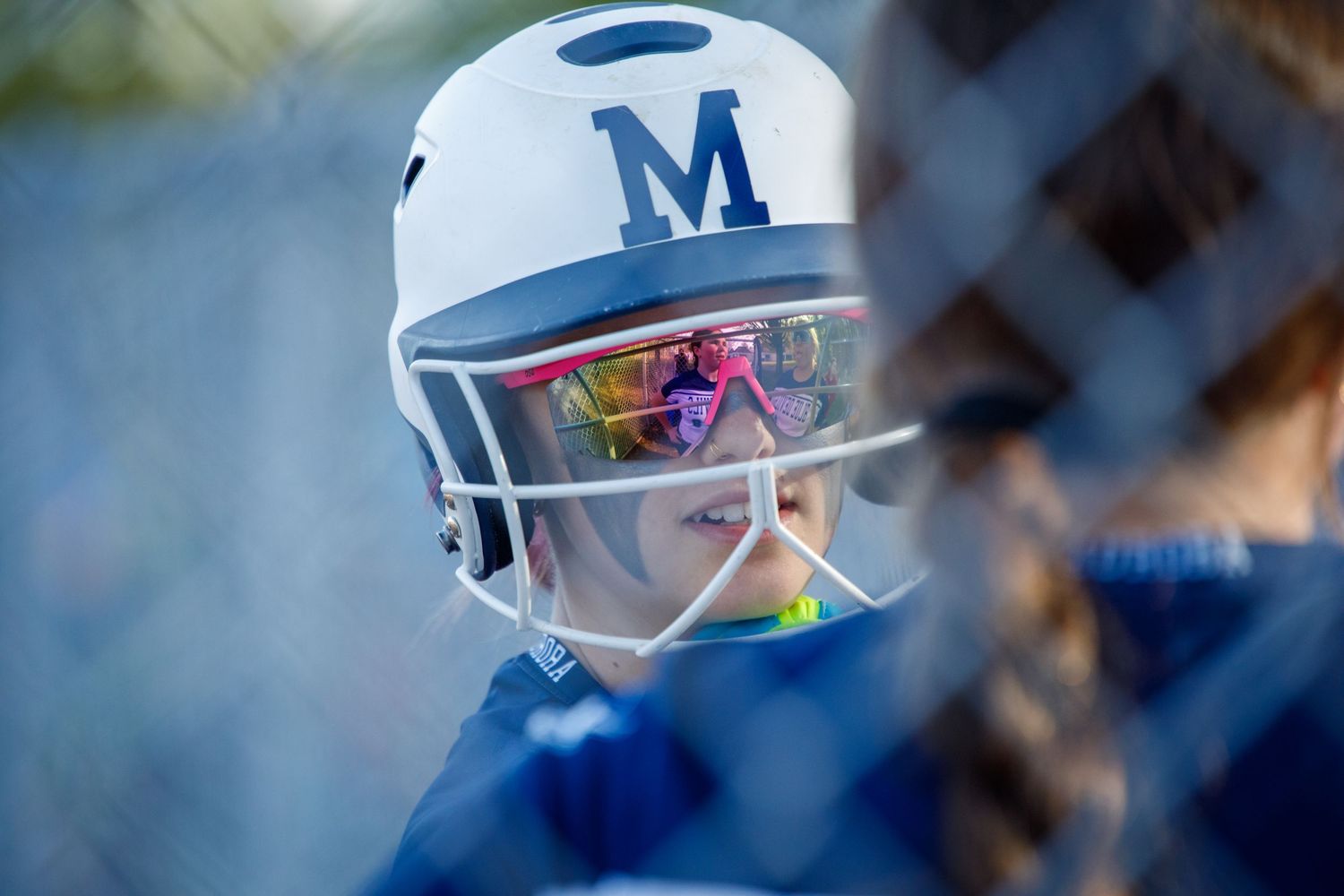 Female softball player wearing white helmet with face guard and blue M logo.