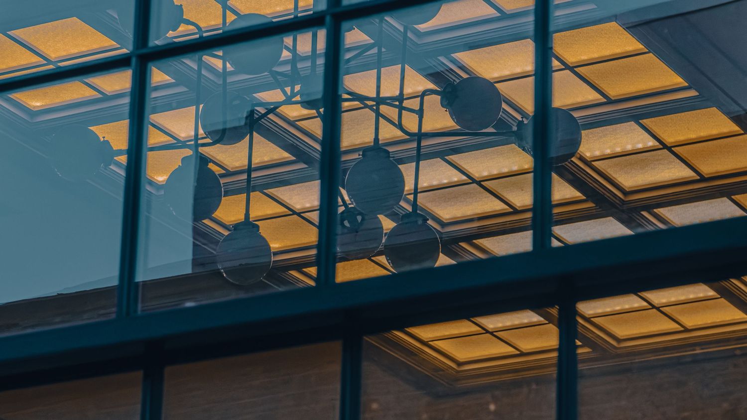 Silhouetted pendant lights reflected in glass building facade with warm golden ceiling panels.