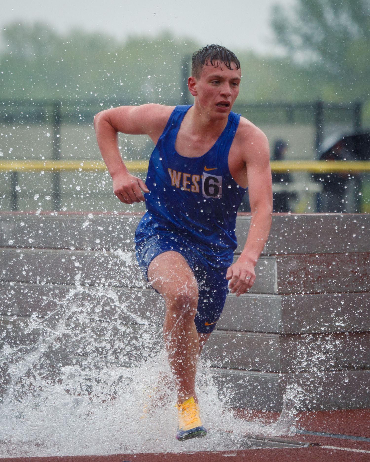 High school track athlete in blue West uniform runs through the water jump obstacle in a steeplechase event.