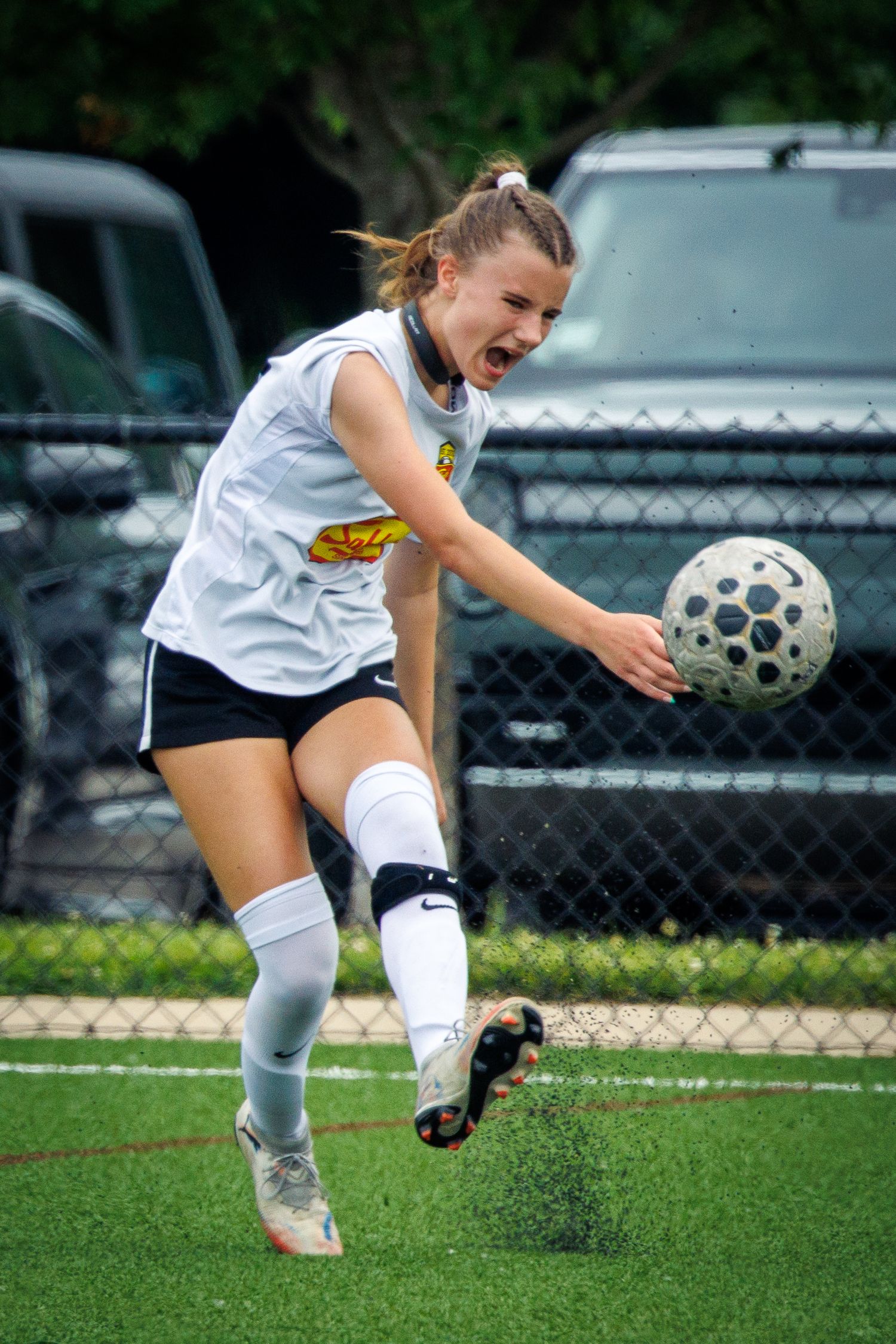 A female soccer player in white kicks the ball with intensity on a turf field during a game.