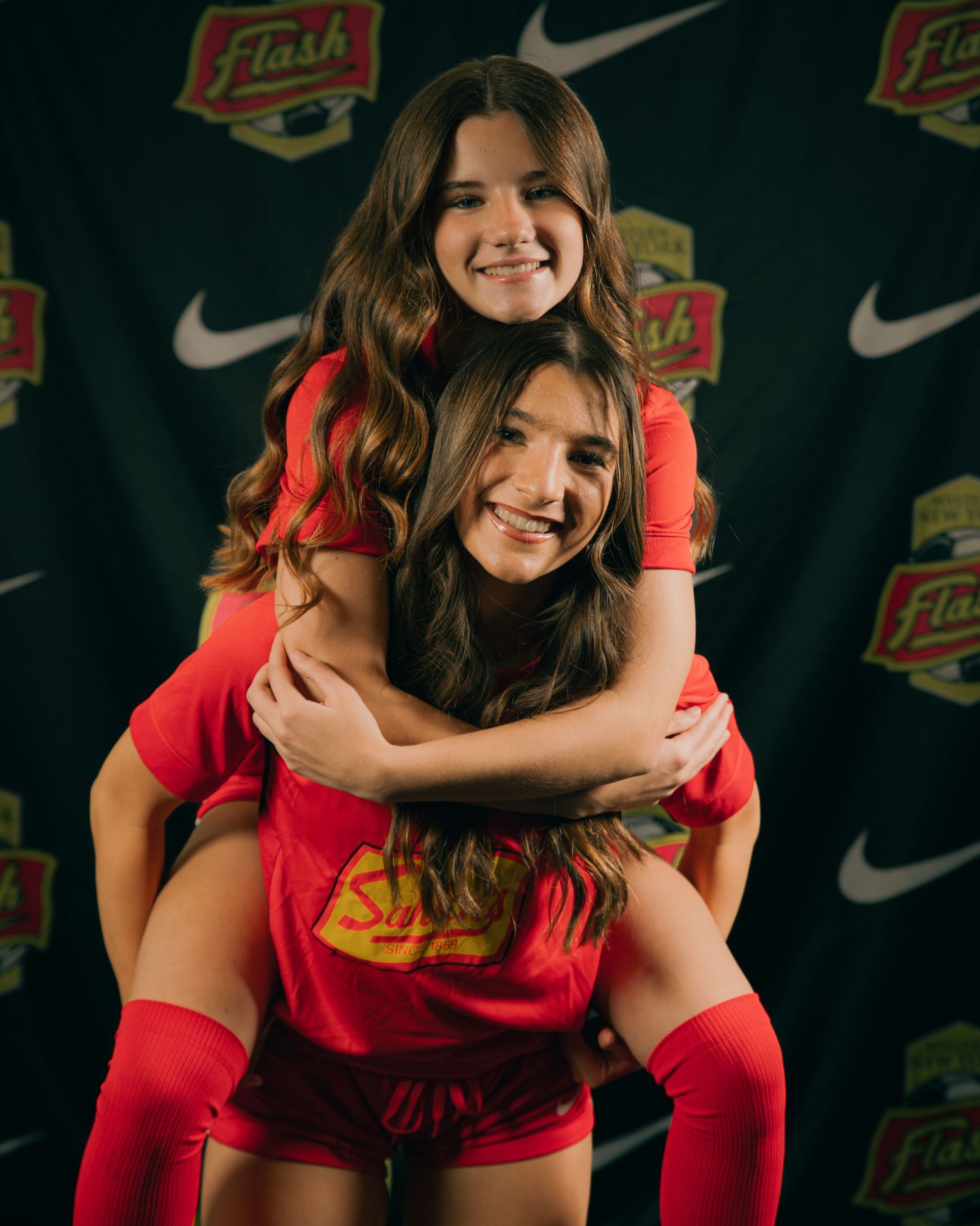 Two female soccer players in red Flash uniforms smiling, one giving the other a piggyback ride.