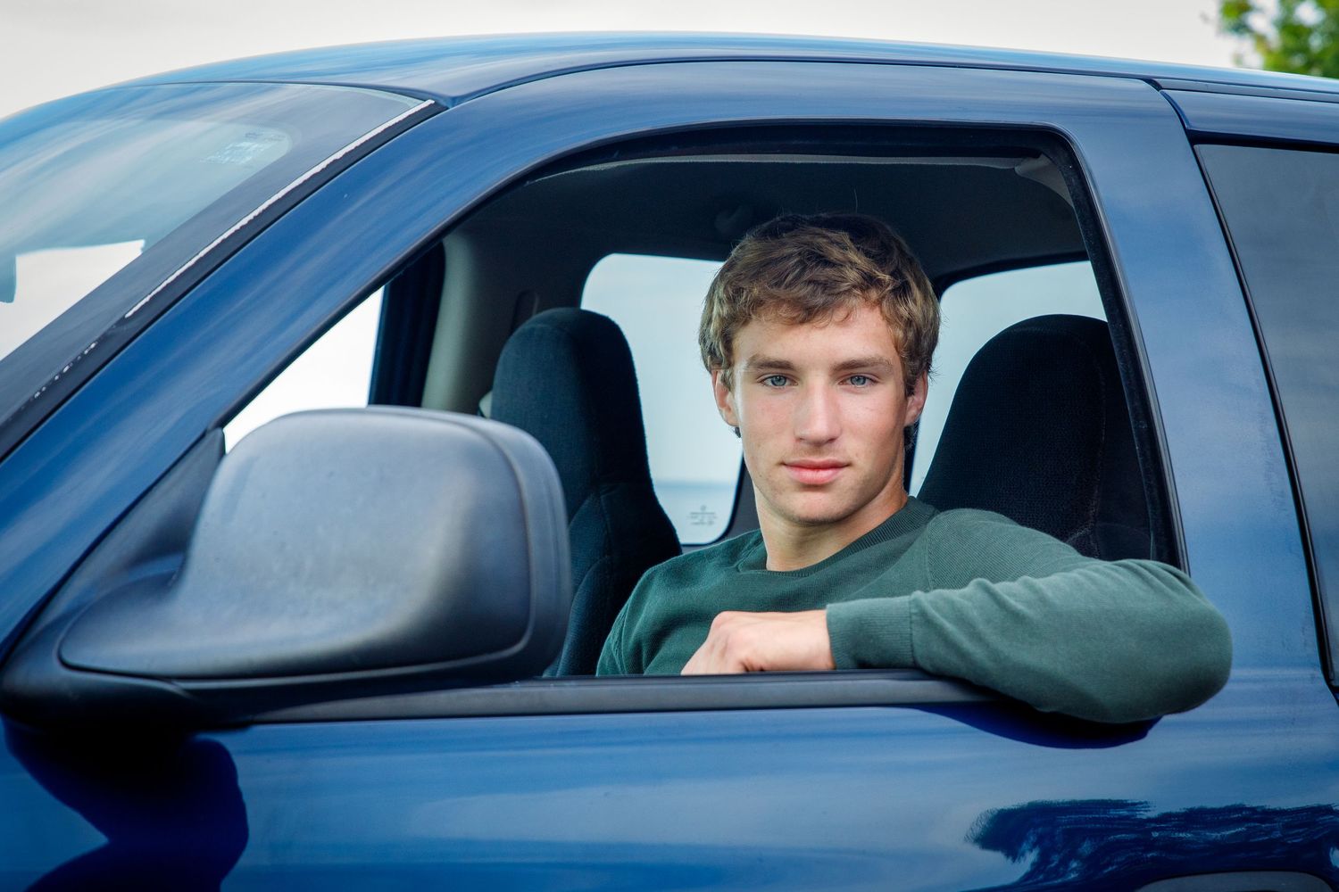 Young man in green shirt sitting in driver seat of blue truck, leaning arm on window.