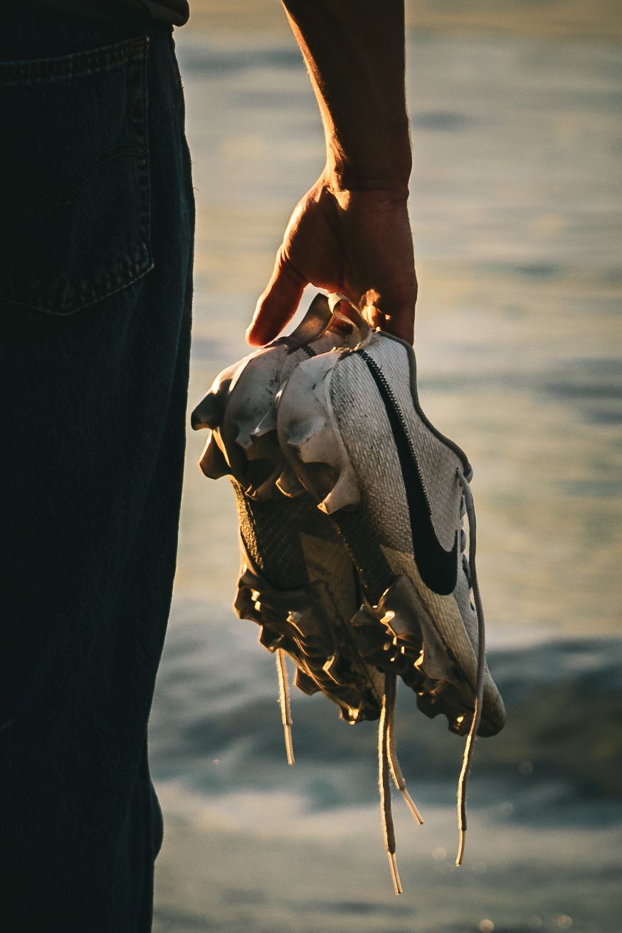 Person holding white Nike sneakers with untied laces by the ocean at golden hour sunset.