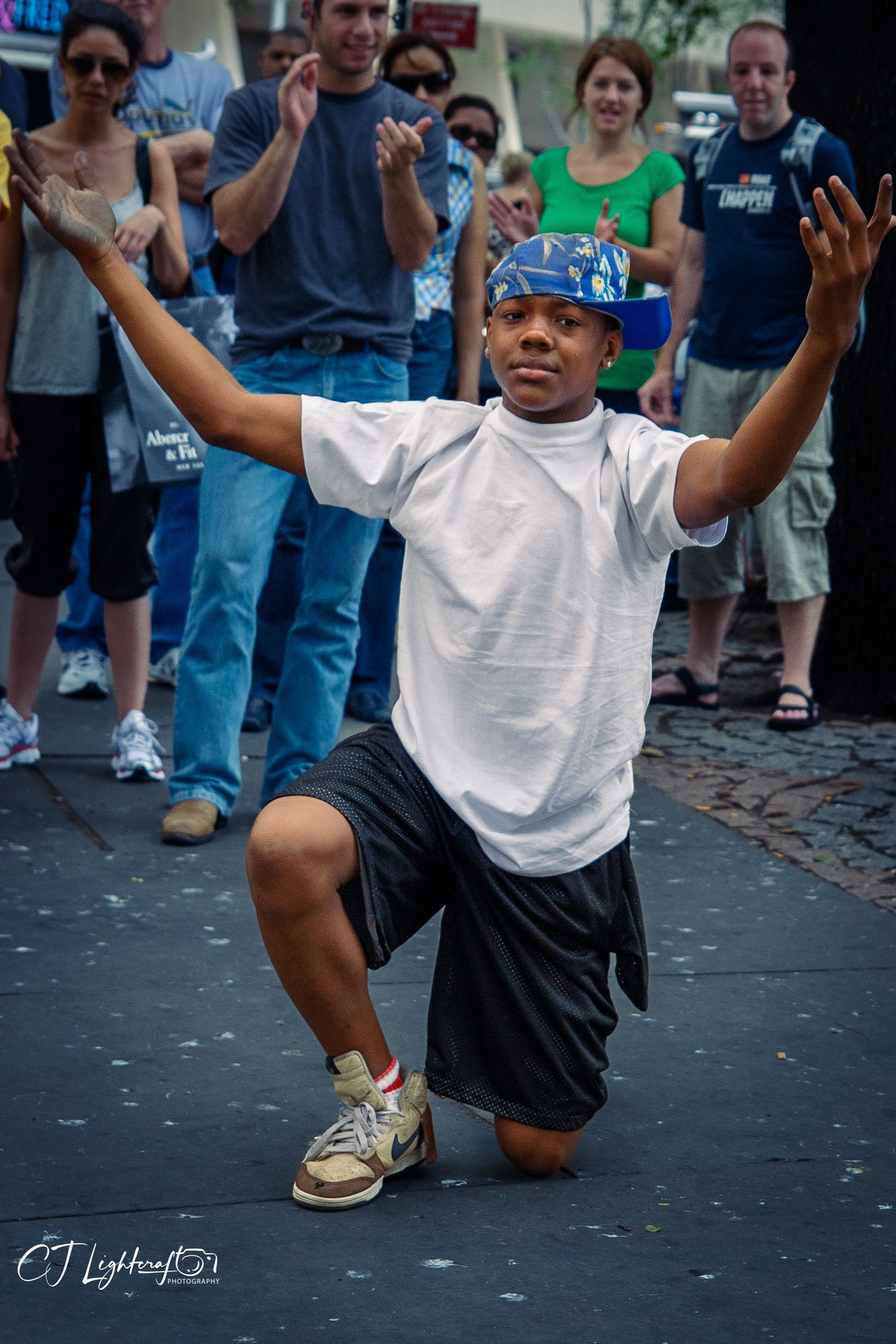 A young break dancer performs a low crouch move for a crowd on a cobblestone street.