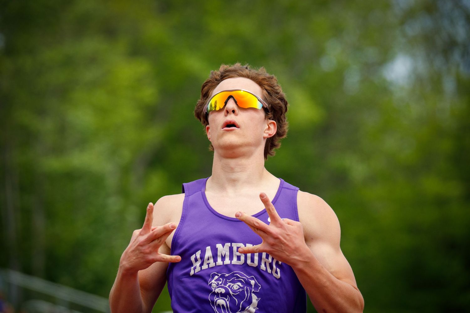 Male track athlete in purple Hamburg jersey makes finger guns gesture wearing orange sport sunglasses.