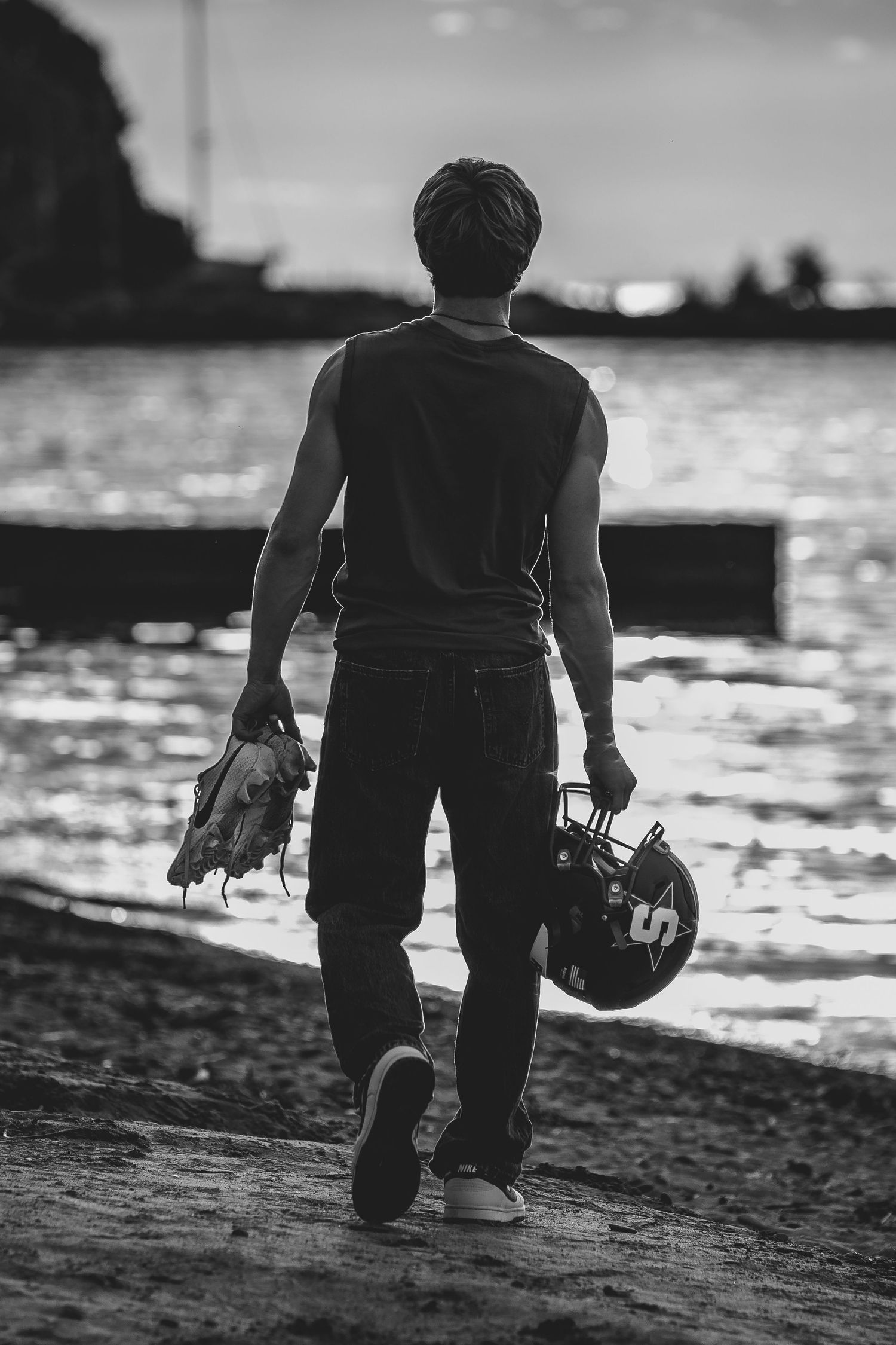 Black and white photo of a man walking by water holding a baseball glove and helmet.