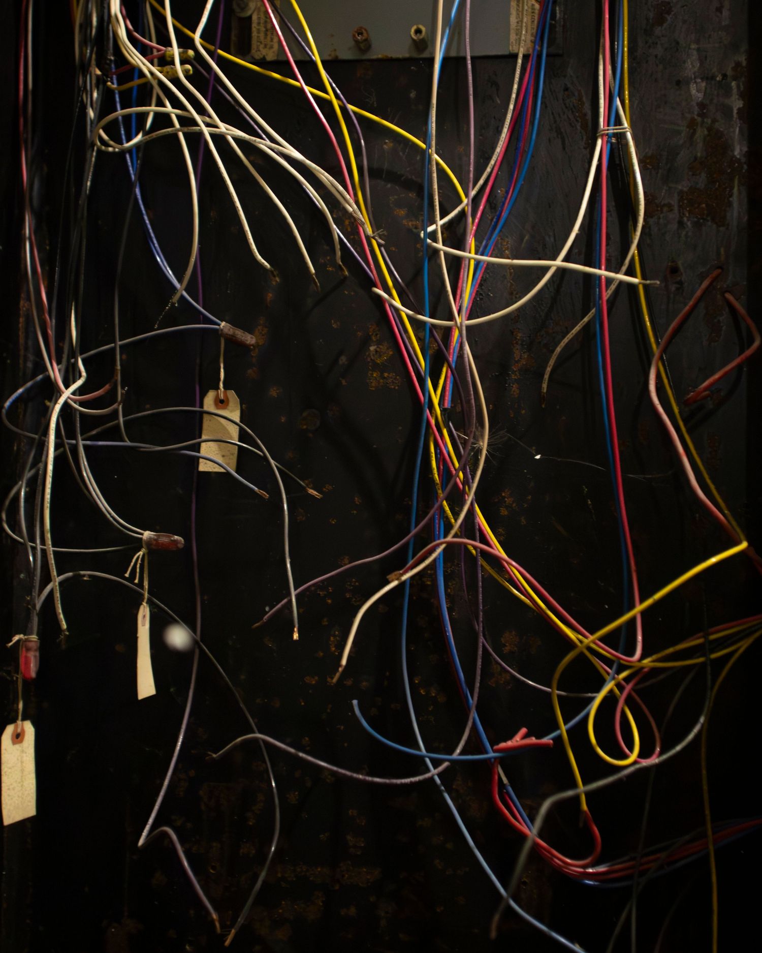 Tangled colorful electrical wires with hanging tags against a dark rusty metal wall panel.