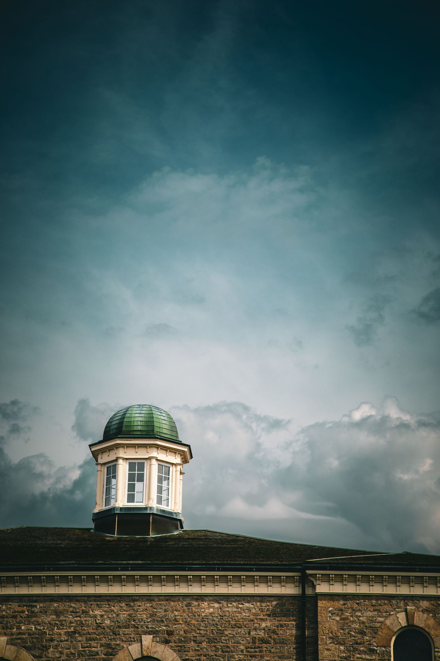 Historic stone building with green domed cupola against dramatic cloudy blue sky backdrop.