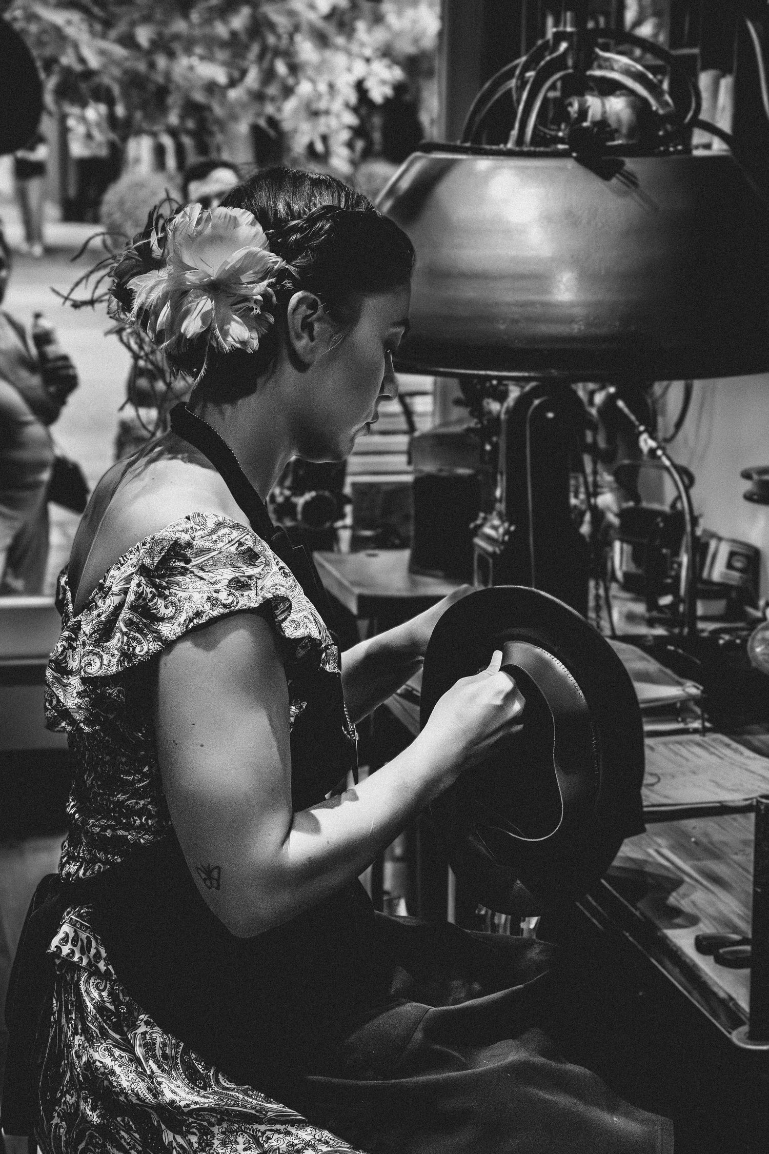 A woman in a floral dress shapes a felt hat using vintage millinery equipment at an outdoor market.