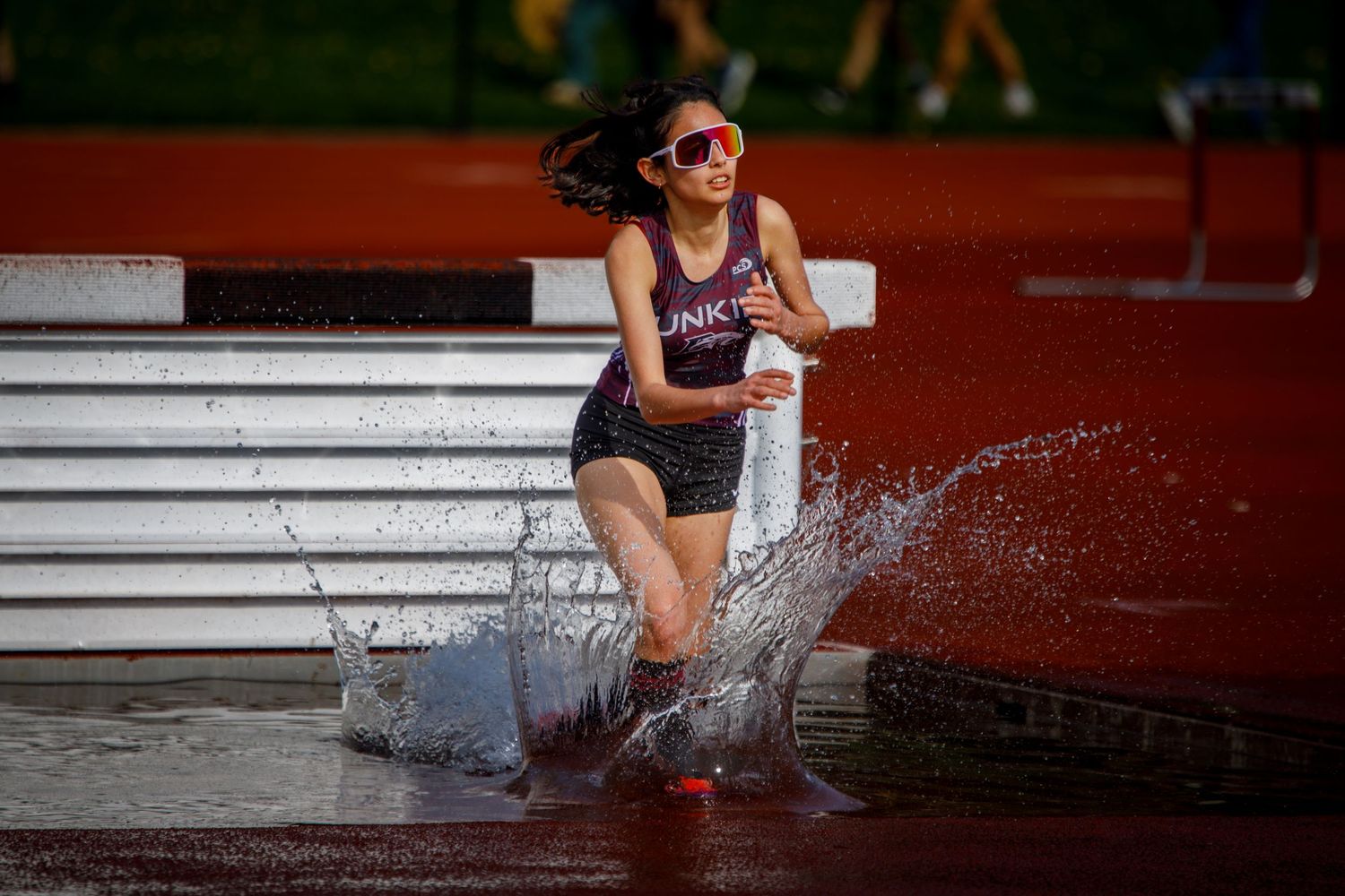 A female runner splashes through a water obstacle during a steeplechase race on a red track.