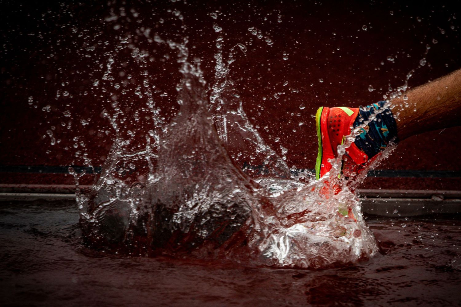 A red cricket ball splashing through water with dramatic impact spray against a dark red background.