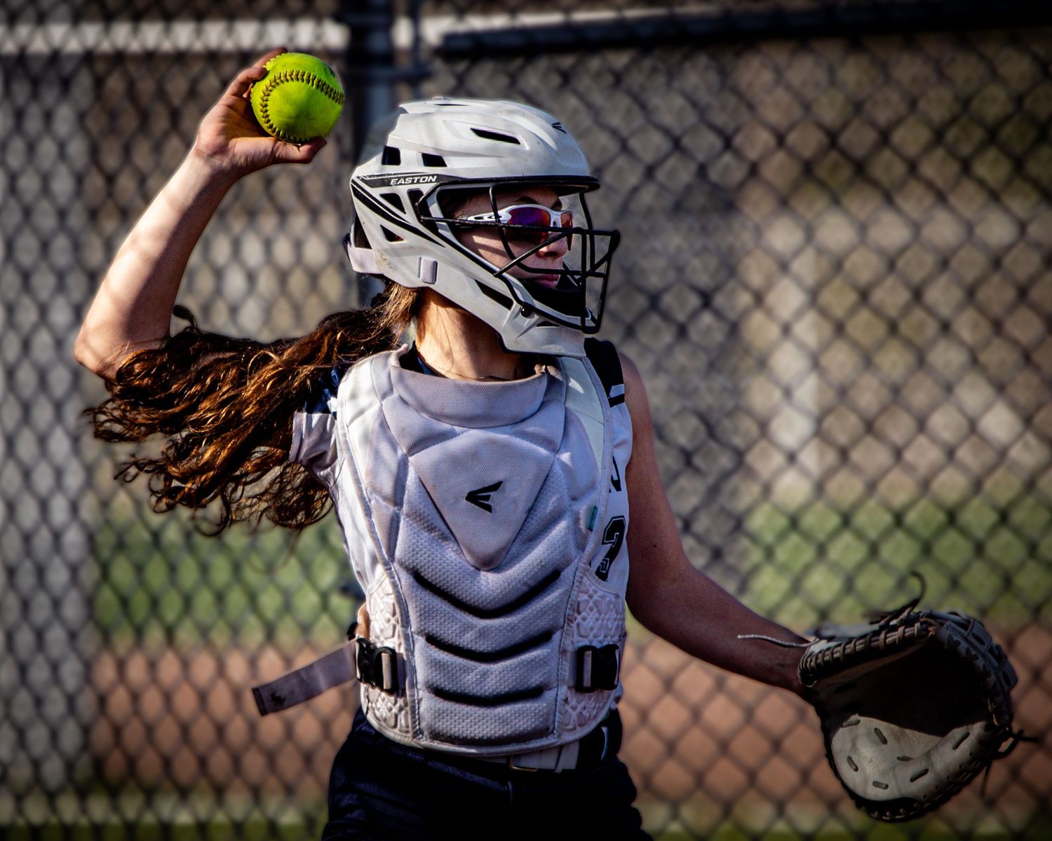 Female softball catcher in Easton gear throwing yellow ball with hair flowing.