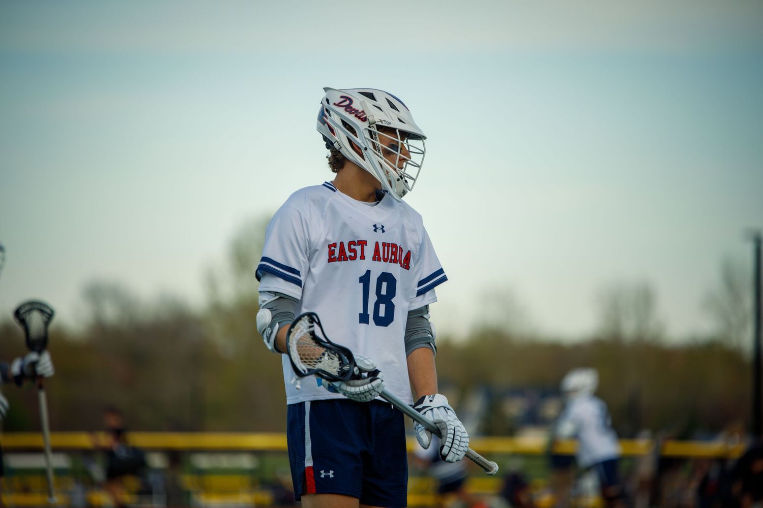 East Aurora lacrosse player #18 in white jersey standing on outdoor field.