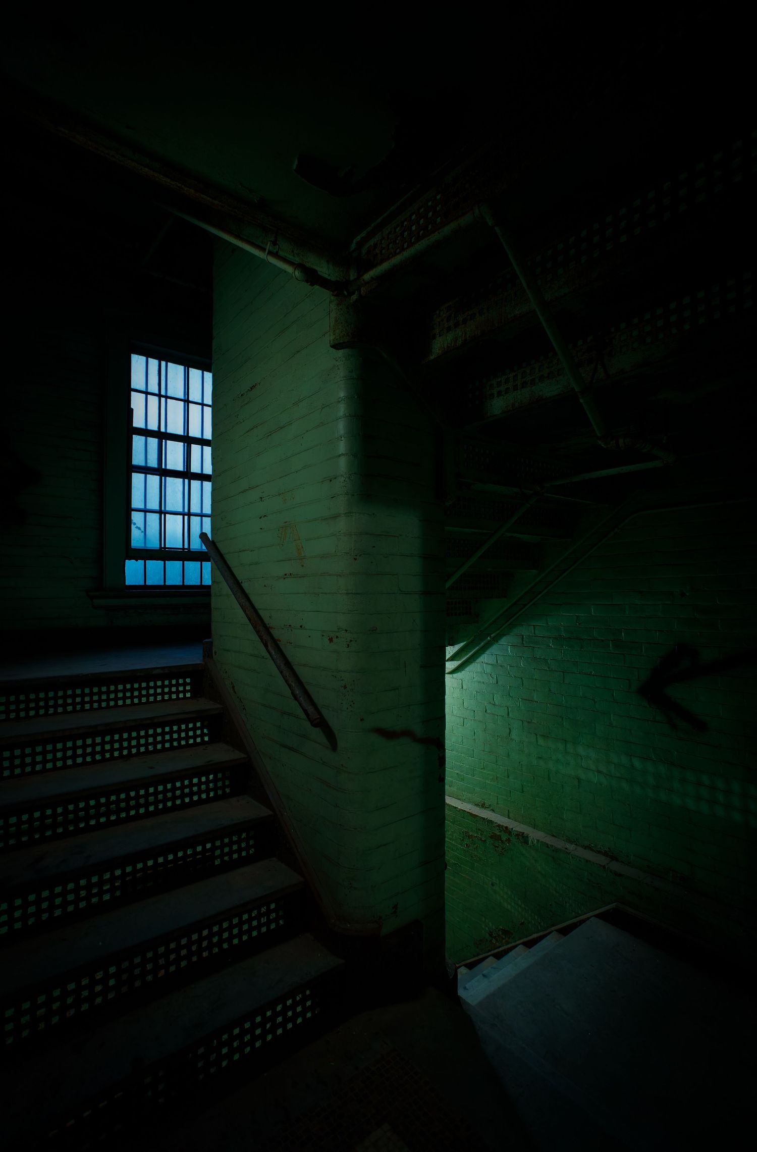 Dark abandoned stairwell with green brick walls and blue light filtering through a barred window.