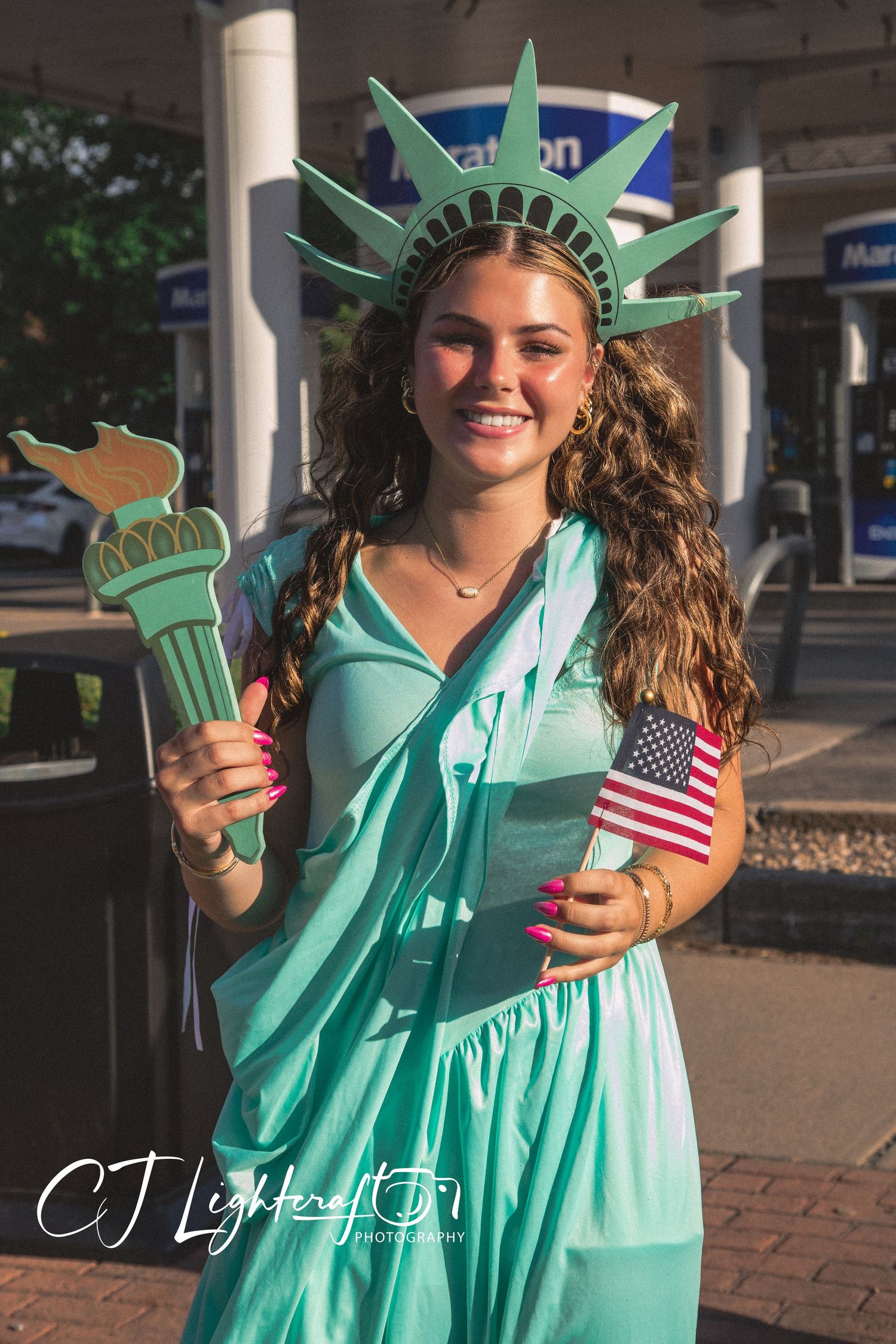 A smiling woman dressed as the Statue of Liberty holds a torch and American flag outdoors.