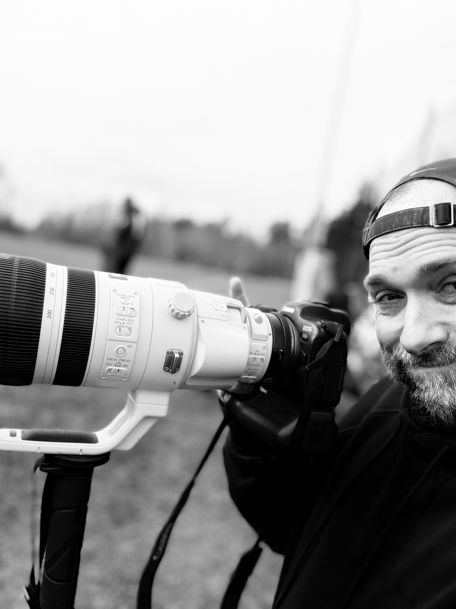 Close-up black and white portrait of a bearded photographer shooting with a large white telephoto lens outdoors.