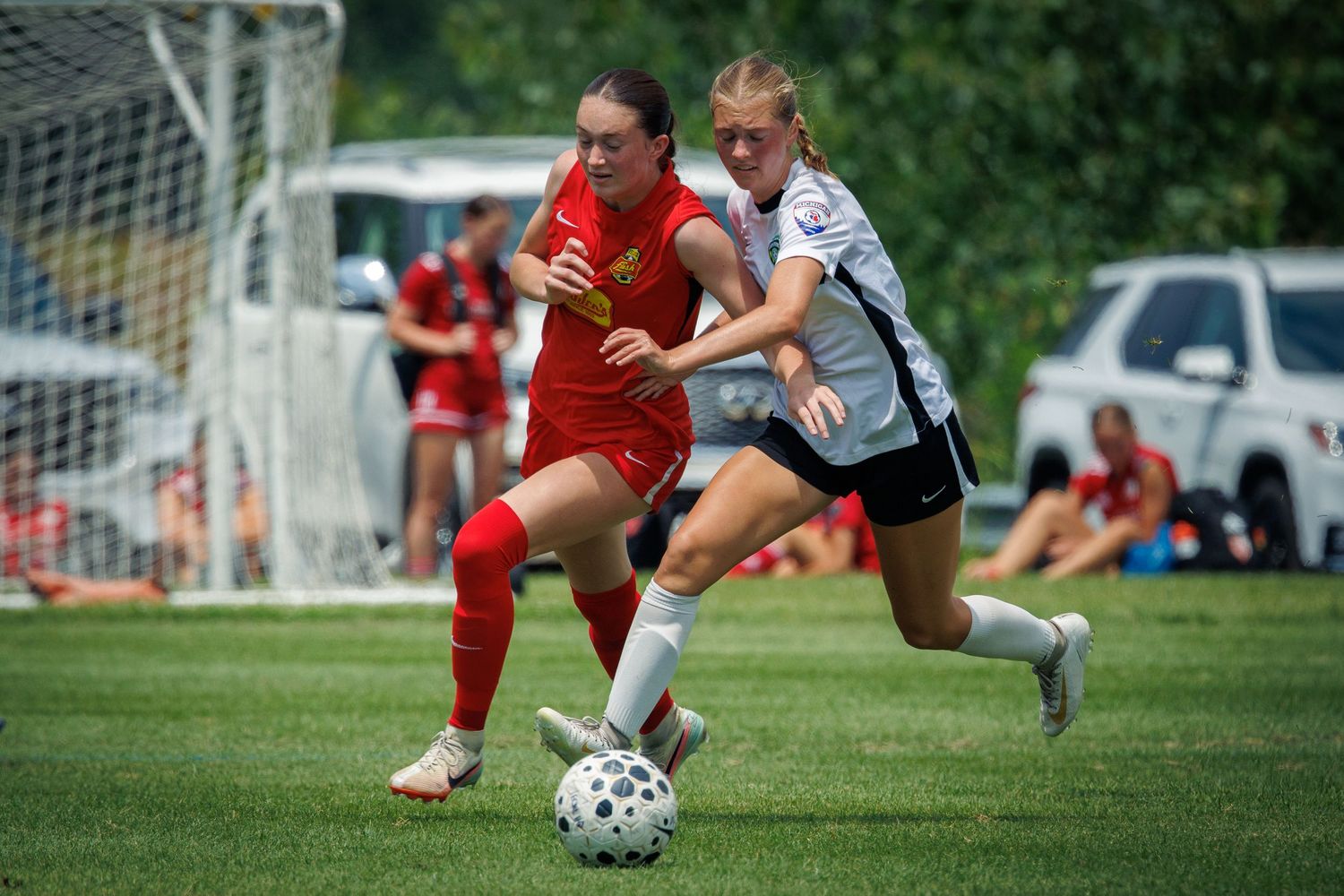 Two girls in red and white uniforms battle for possession of the soccer ball during an outdoor youth soccer game.
