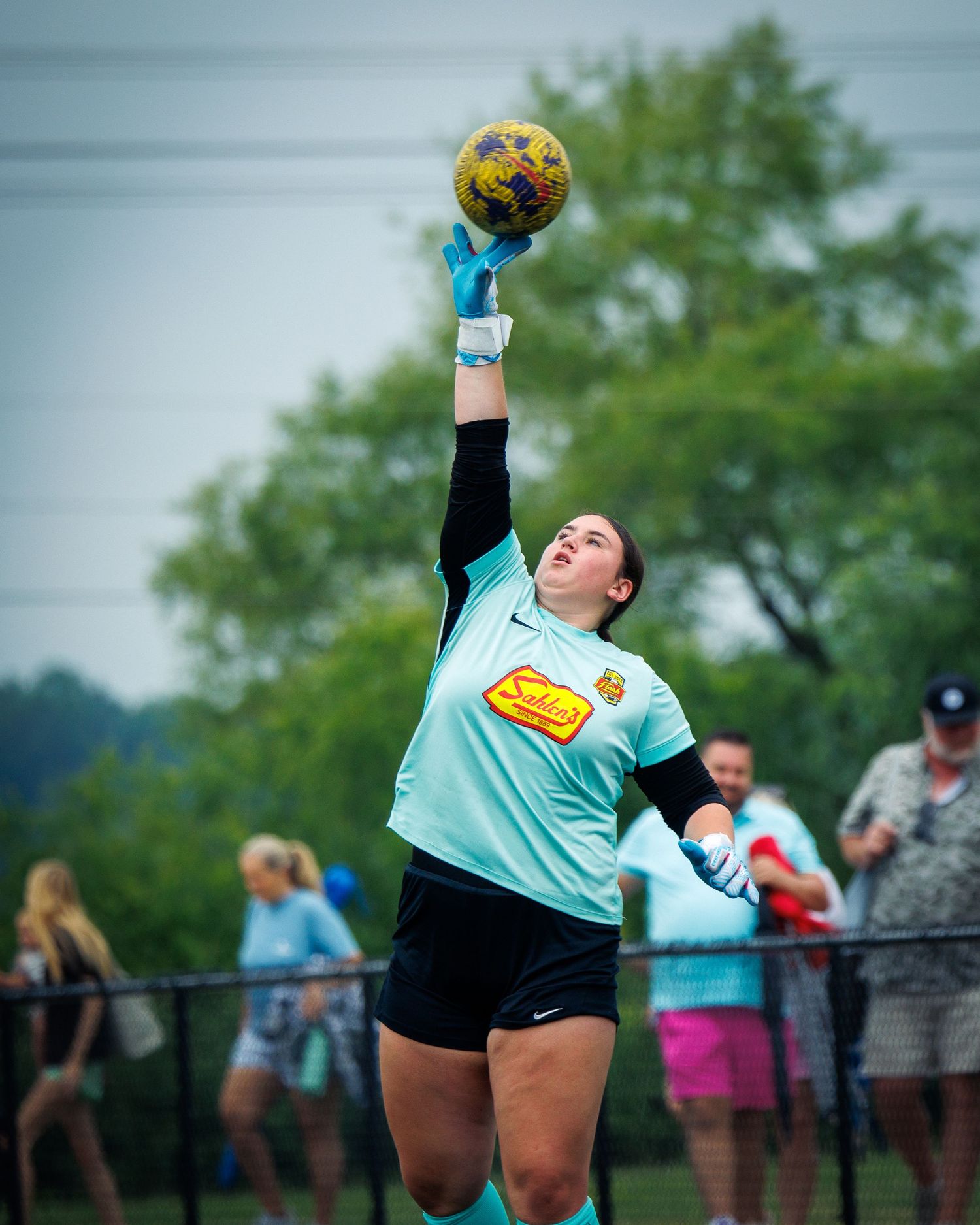 A soccer goalkeeper in a teal jersey reaches up to catch a colorful ball during an outdoor match.